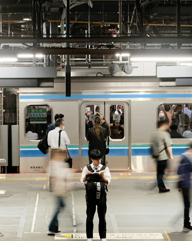 Commuters at a busy train station platform, with a train arriving in the background. People are walking, and one person is standing still, looking at a device.