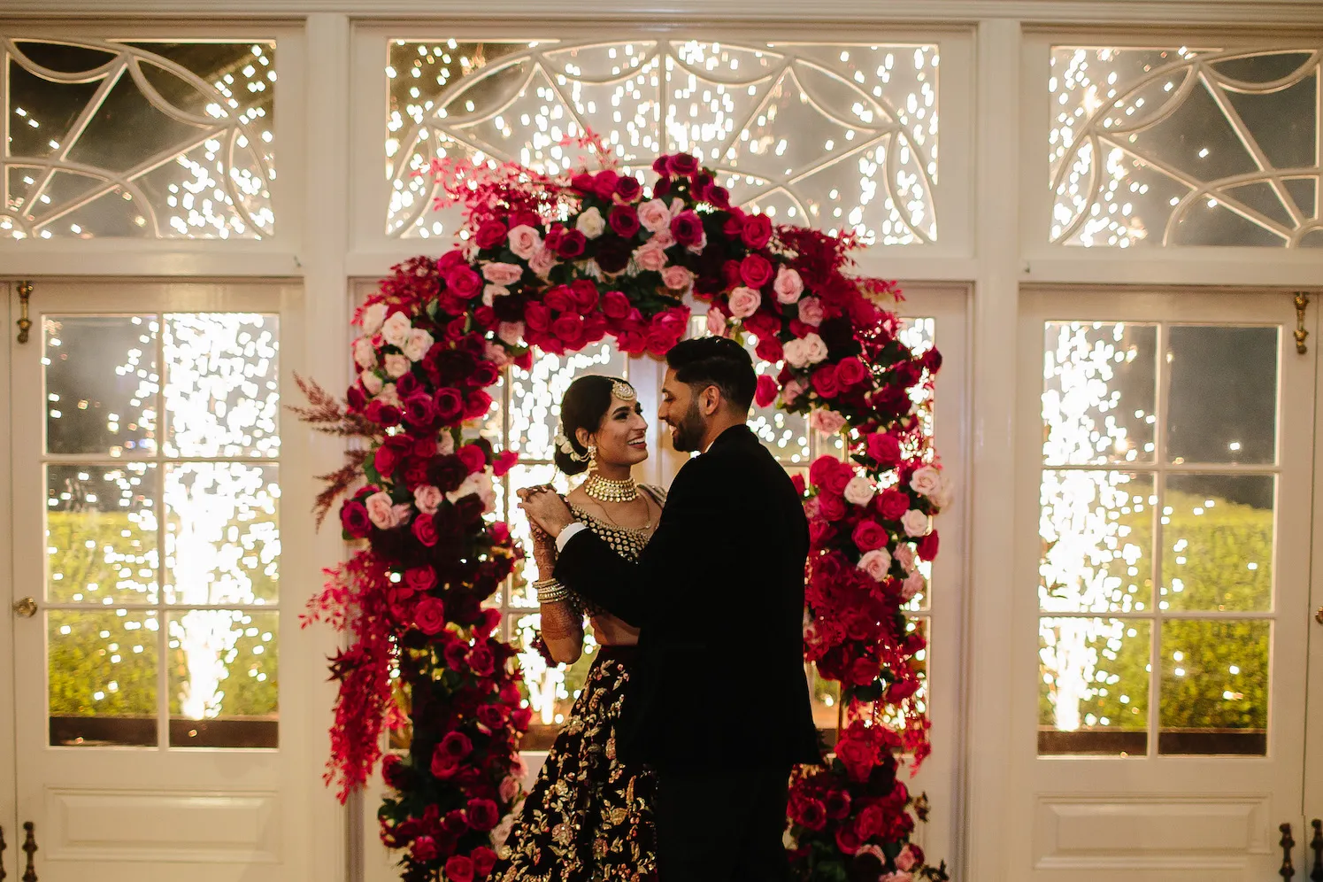 A couple dressed in formal attire stands embraced in front of a beautifully decorated floral arch with red and pink roses. The background features bright indoor fireworks, creating a celebratory and romantic atmosphere.