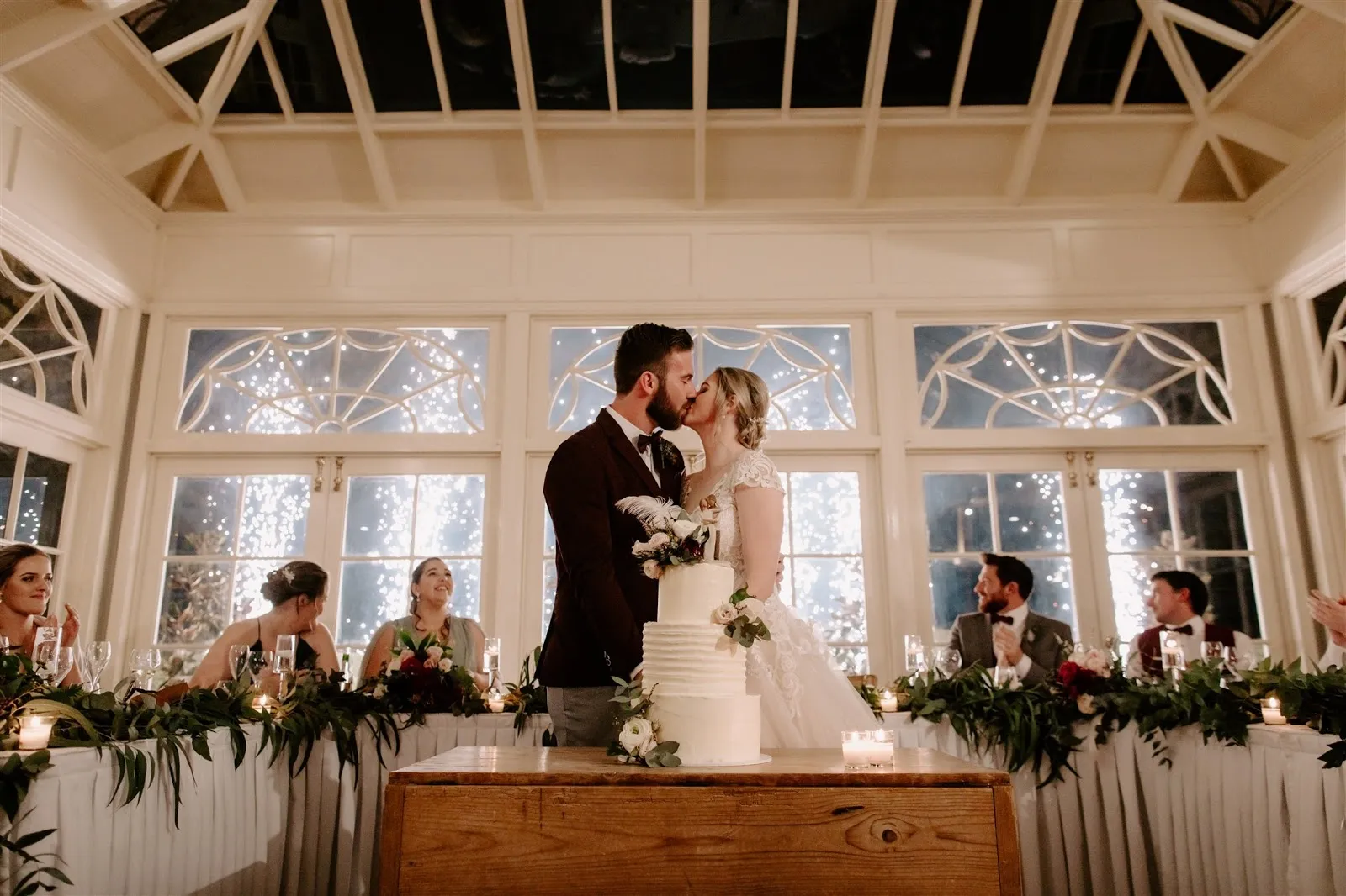 A bride and groom share a kiss behind their wedding cake in a beautifully decorated room with a glass ceiling. Bridesmaids and groomsmen are seated at a table covered with greenery and candles, and twinkling lights illuminate the background.