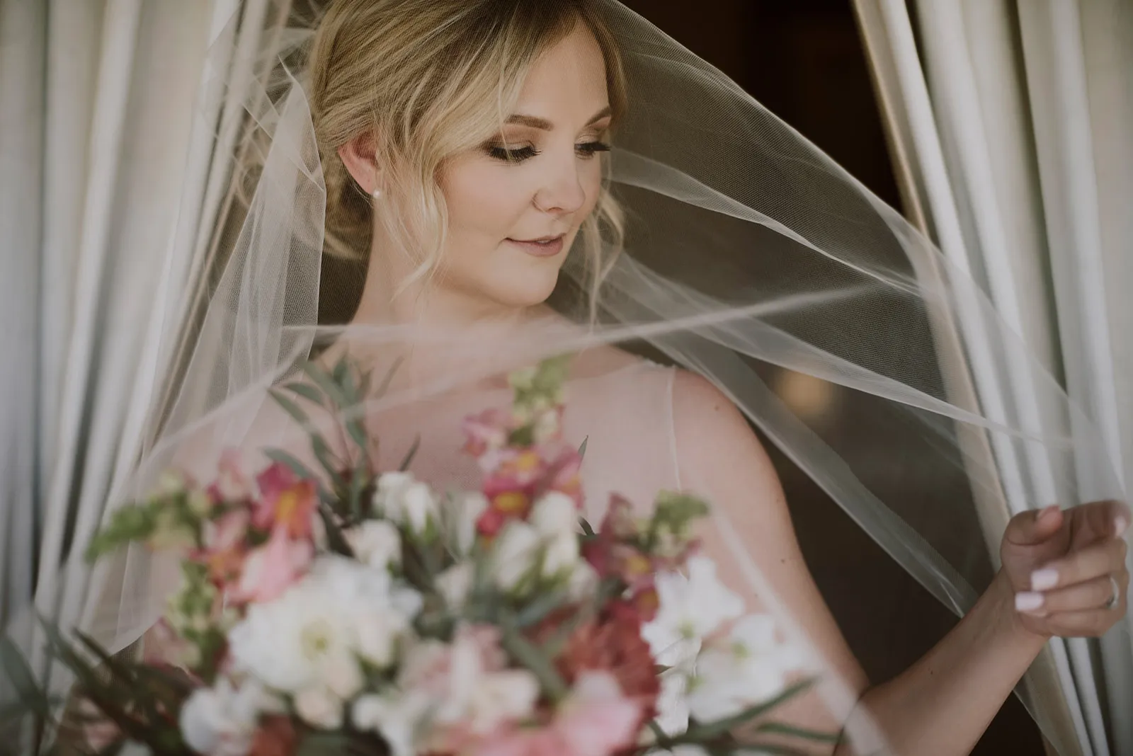 A bride stands near a window, softly lit by natural light. She is looking down and smiling gently, holding a bouquet of pink and white flowers close to her chest. A thin veil drapes over her hair and shoulders, adding an ethereal quality to the scene.
