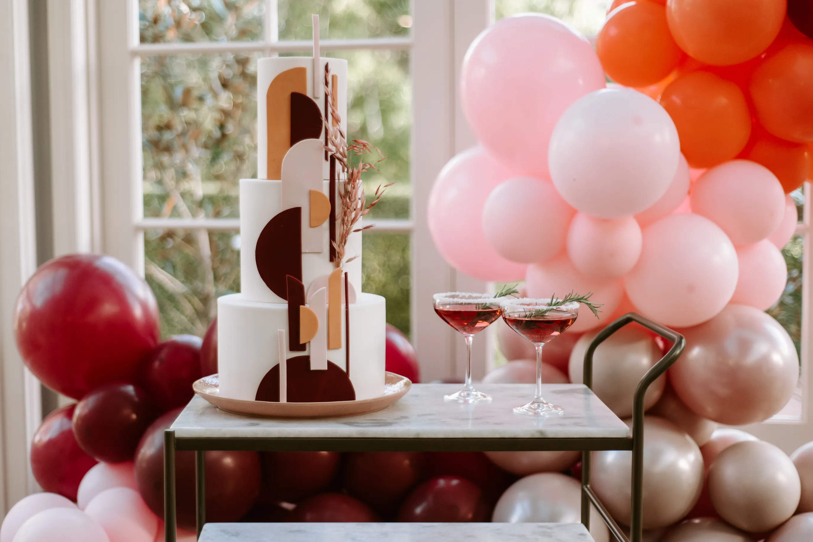 A modern white cake with geometric decorations in shades of brown, yellow, and white is displayed on a marble table. Two cocktail glasses with a reddish drink and garnishes sit beside the cake. The background features large balloons in red, pink, and beige hues.