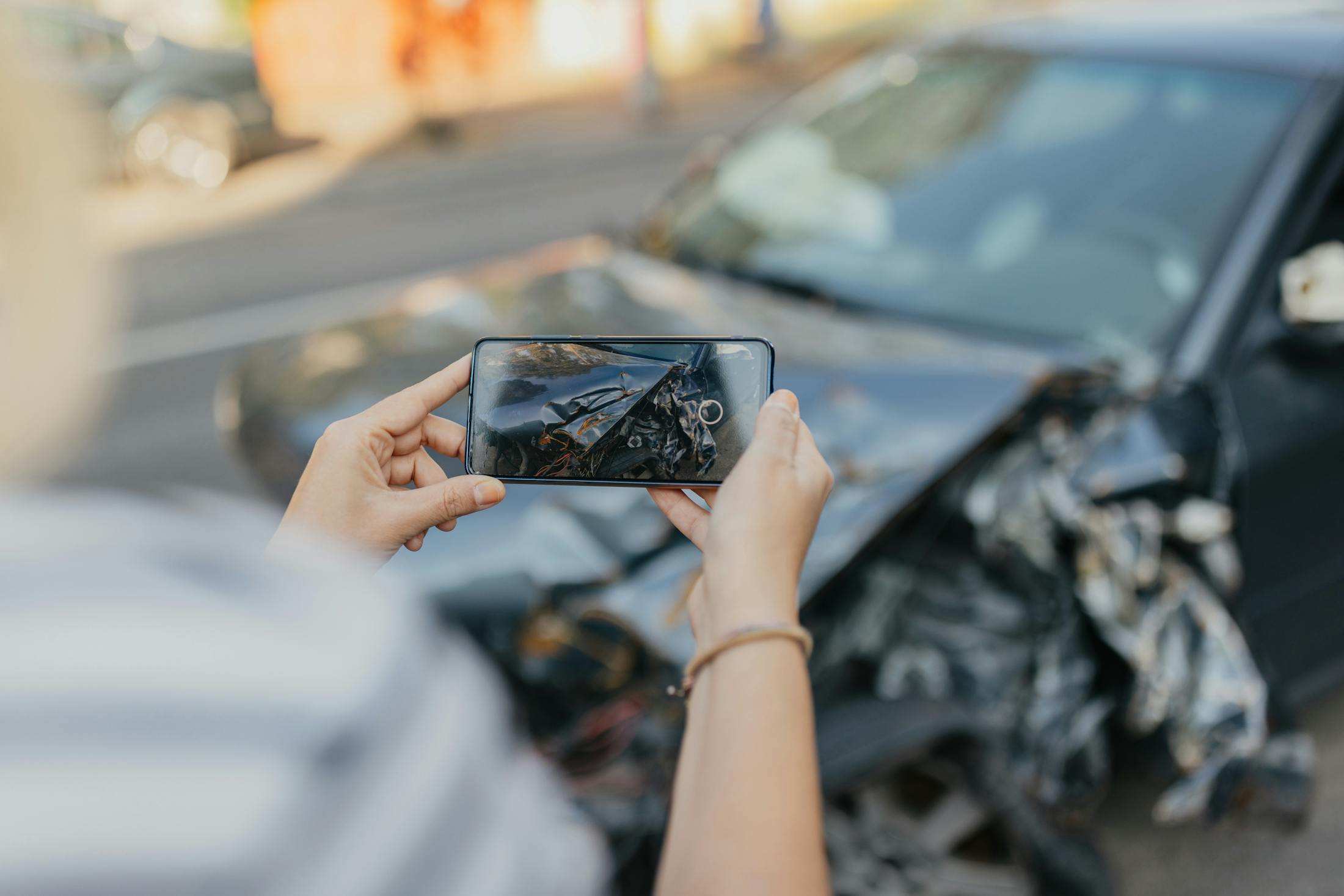A person is taking a photo with their smart phone of a bloack car that has been totaled from a rideshare accident