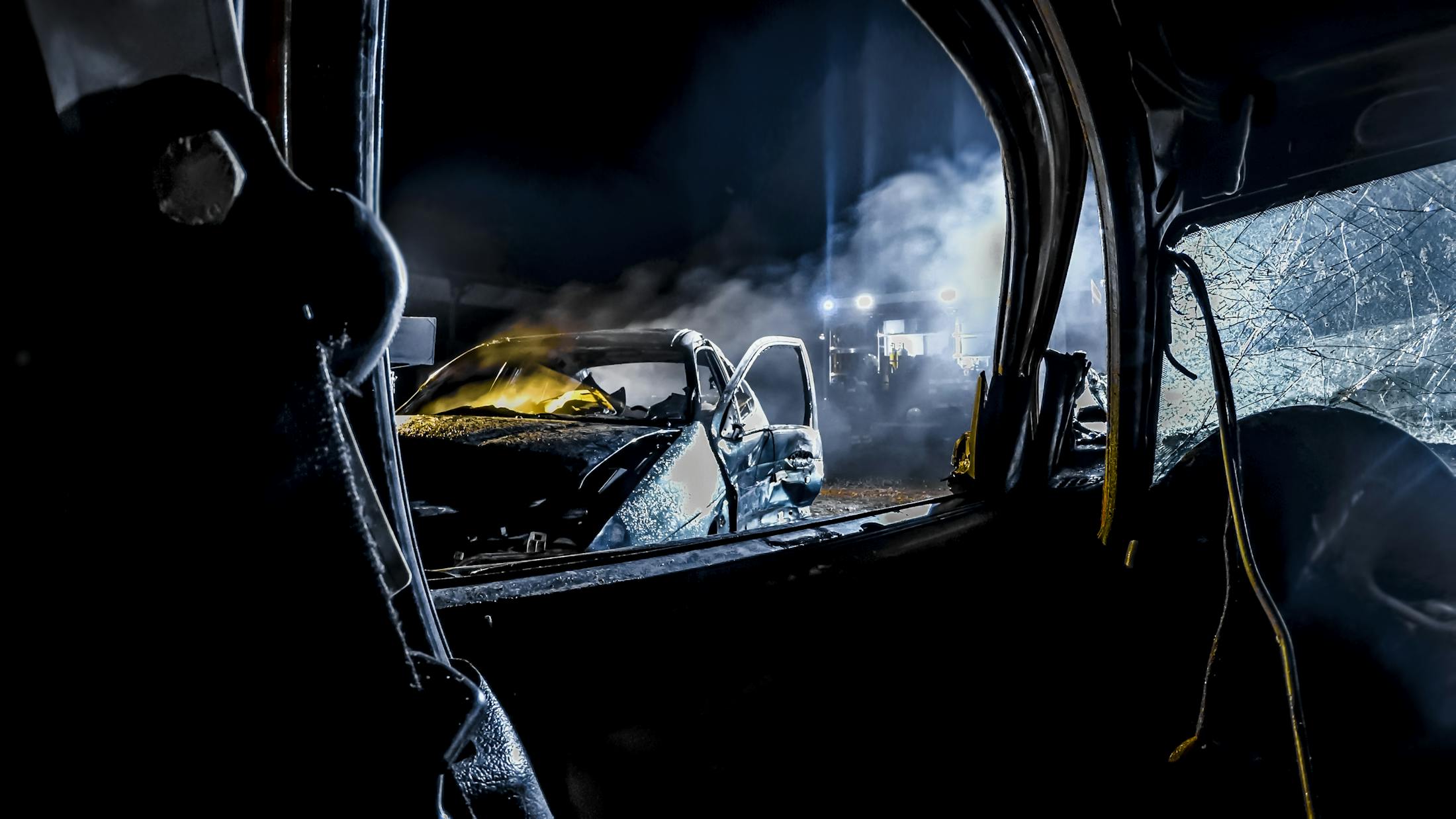 An image of a car accident taken from the backseat of one of the vehicles that has been totaled. It is taken at night through the window from the right backseat through the front driverside window. There is a totaled car with smoke coming from it.