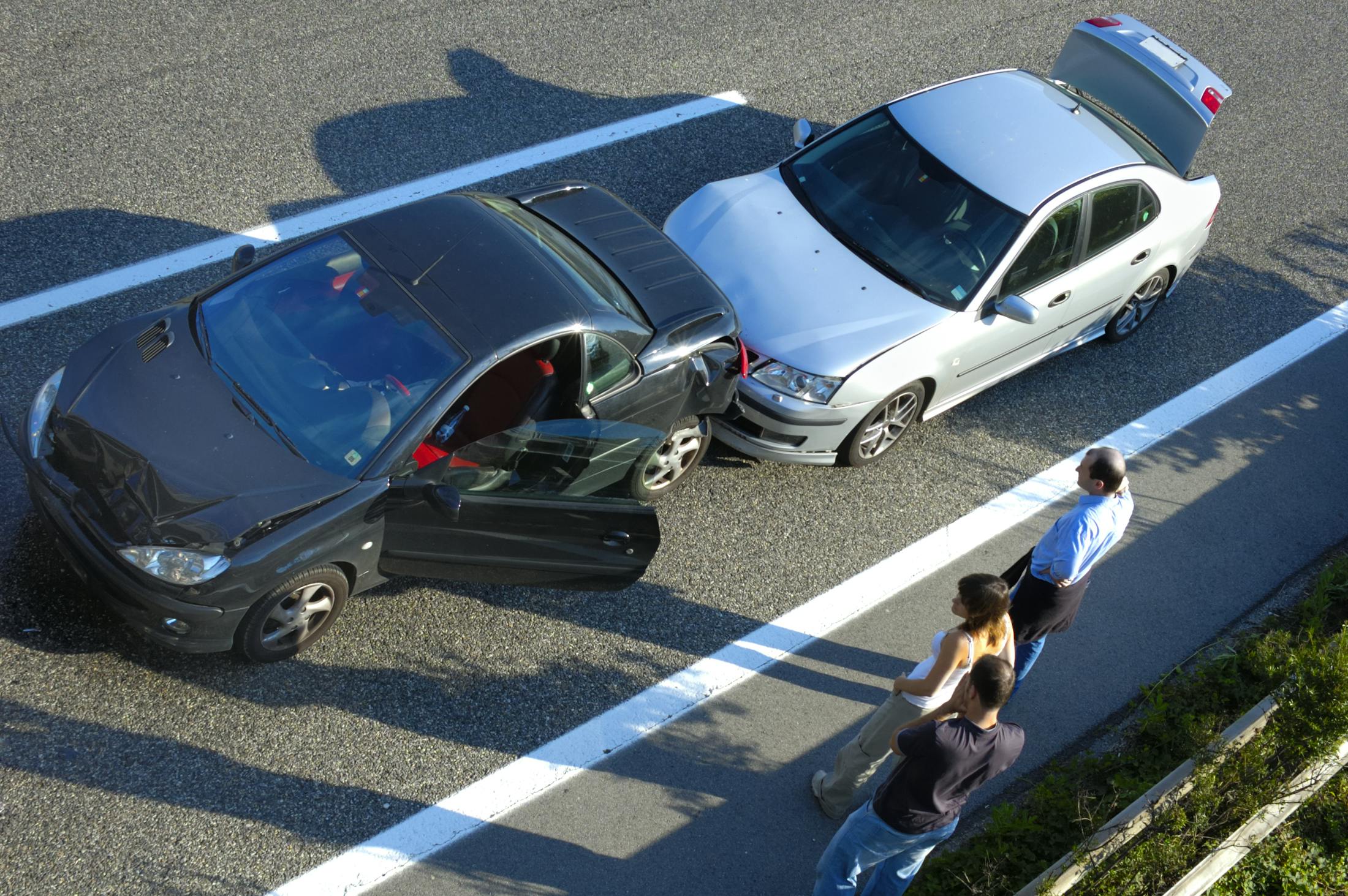 people standing on the side of the road looking at two cars who have been in a fender bender.