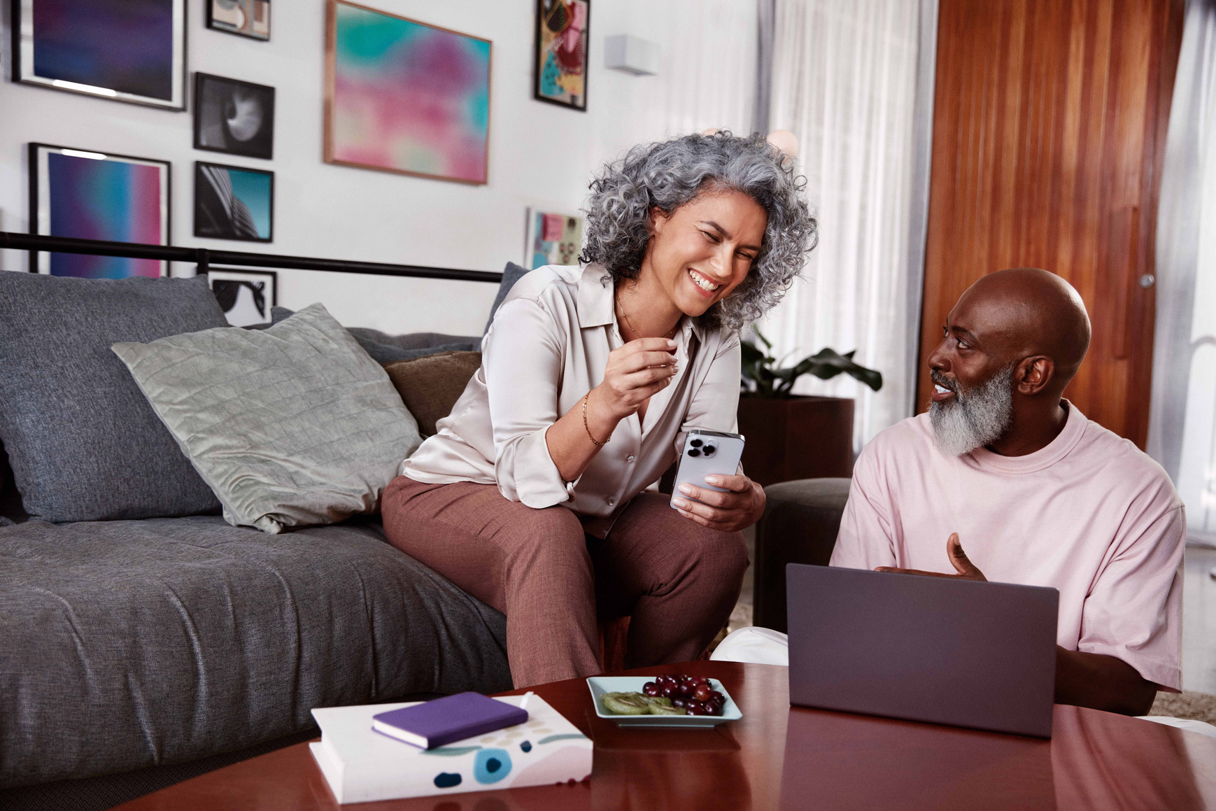 Mulher negra de cabelos cacheados e grisalhos sentada em um sofá e sorrindo, segurando com a mão esquerda um celular em direção a um homem negro, careca, com barba grisalha, sentado ao seu lado no chão, olhando para o rosto dela enquanto sorri e com uma pequena mesa de madeira a sua frente com um notebook.