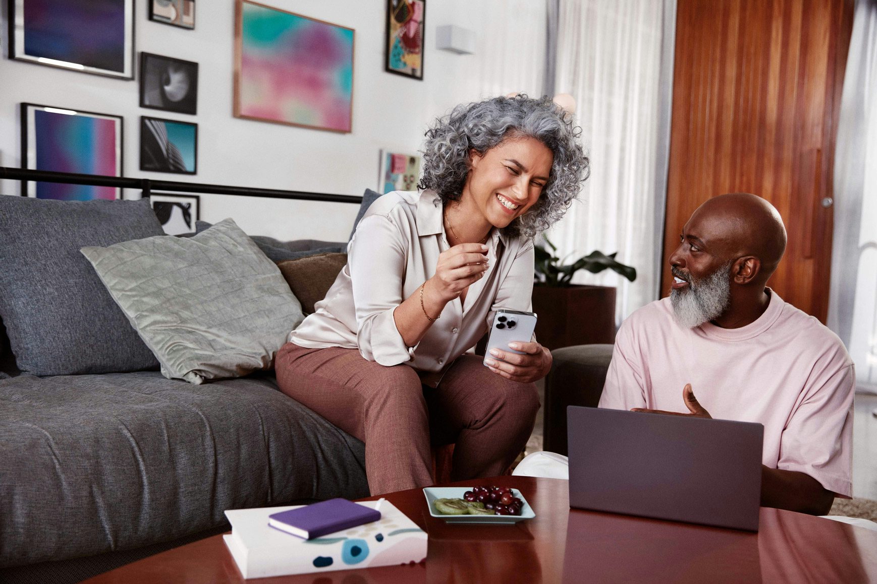 Mulher negra de cabelos cacheados e grisalhos sentada em um sofá e sorrindo, segurando com a mão esquerda um celular em direção a um homem negro, careca, com barba grisalha, sentado ao seu lado no chão, olhando para o rosto dela enquanto sorri e com uma pequena mesa de madeira a sua frente com um notebook.