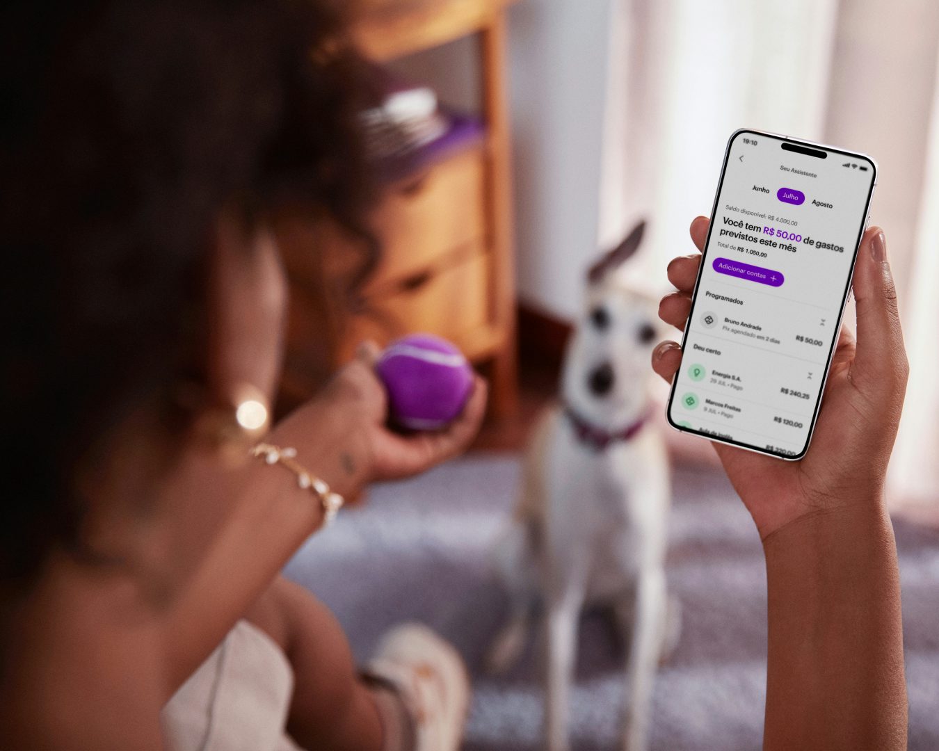 Vista das costas de uma mulher negra sentada, com um celular na mão direita e visualizando sua conta no aplicativo do Nubank, com sua mão esquerda segura uma bolinha de tênis roxa, ao fundo e desfocado um cachorro de pequeno porte sentado no chão olhando para a bolinha