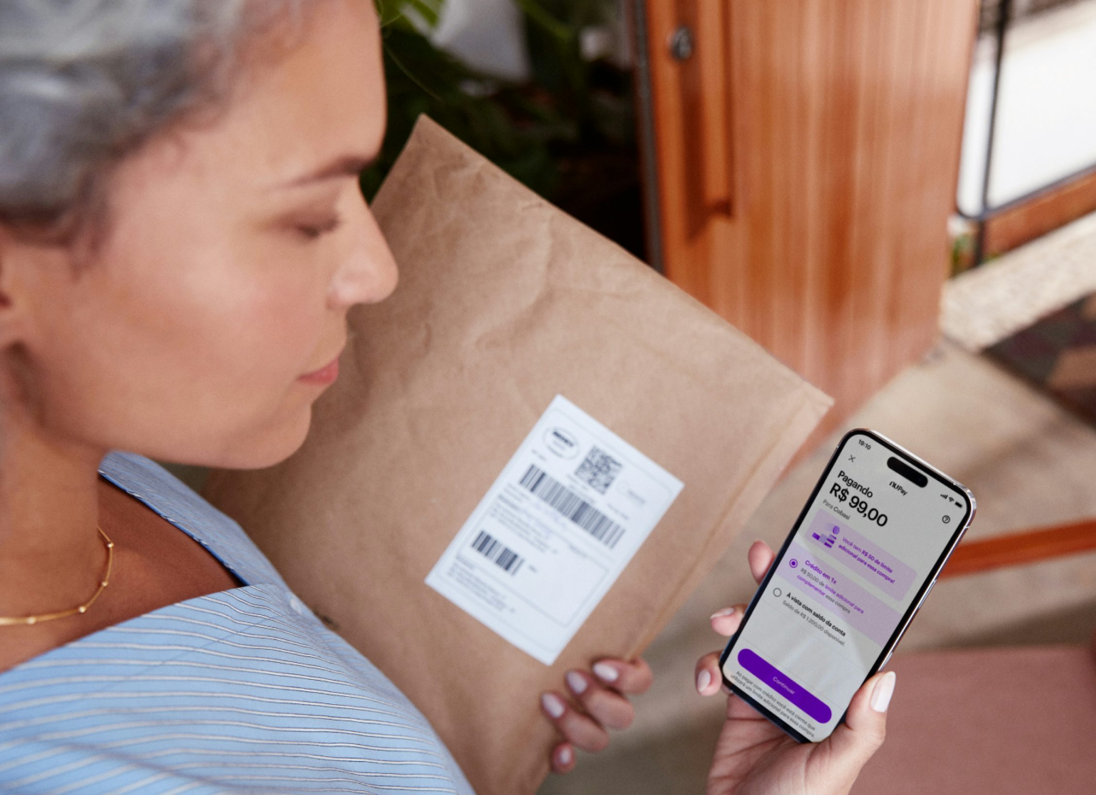 Mulher de blusa azul claro com listras brancas com a mão esquerda segurando um envelope e na mão direita segurando o celular com o app Nubank na tela mostrando uma transação de pagamento.