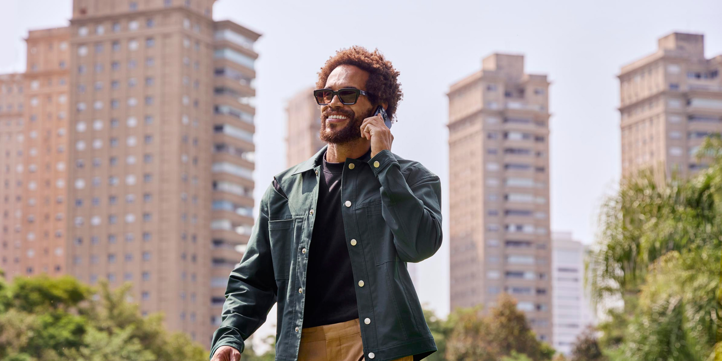 Homem sorrindo enquanto fala no celular passeando pela cidade. Ele está segurando o celular com a mão esquerda e, ao fundo, prédios e algumas árvores.
