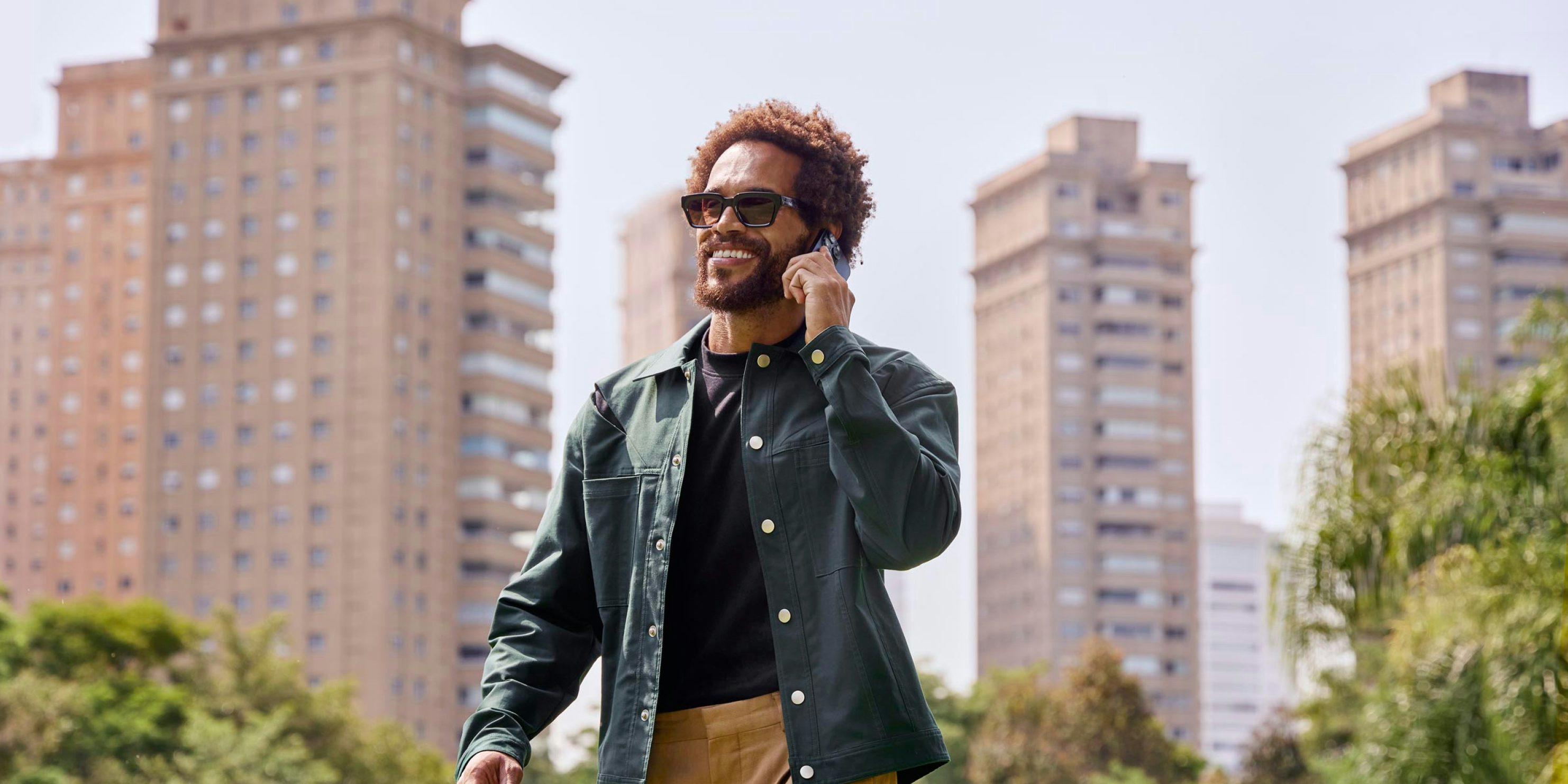Homem sorrindo enquanto fala no celular passeando pela cidade. Ele está segurando o celular com a mão esquerda e, ao fundo, prédios e algumas árvores.