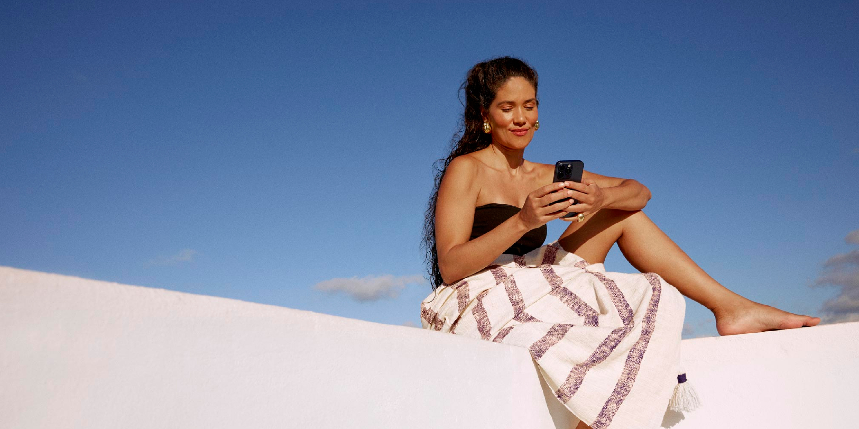 Mulher de top e uma canga sobre as pernas está sentada em uma superfície branca na luz do dia com o céu azul. Ela segura o celular com as duas mãos e olha para a tela com um sorriso leve.