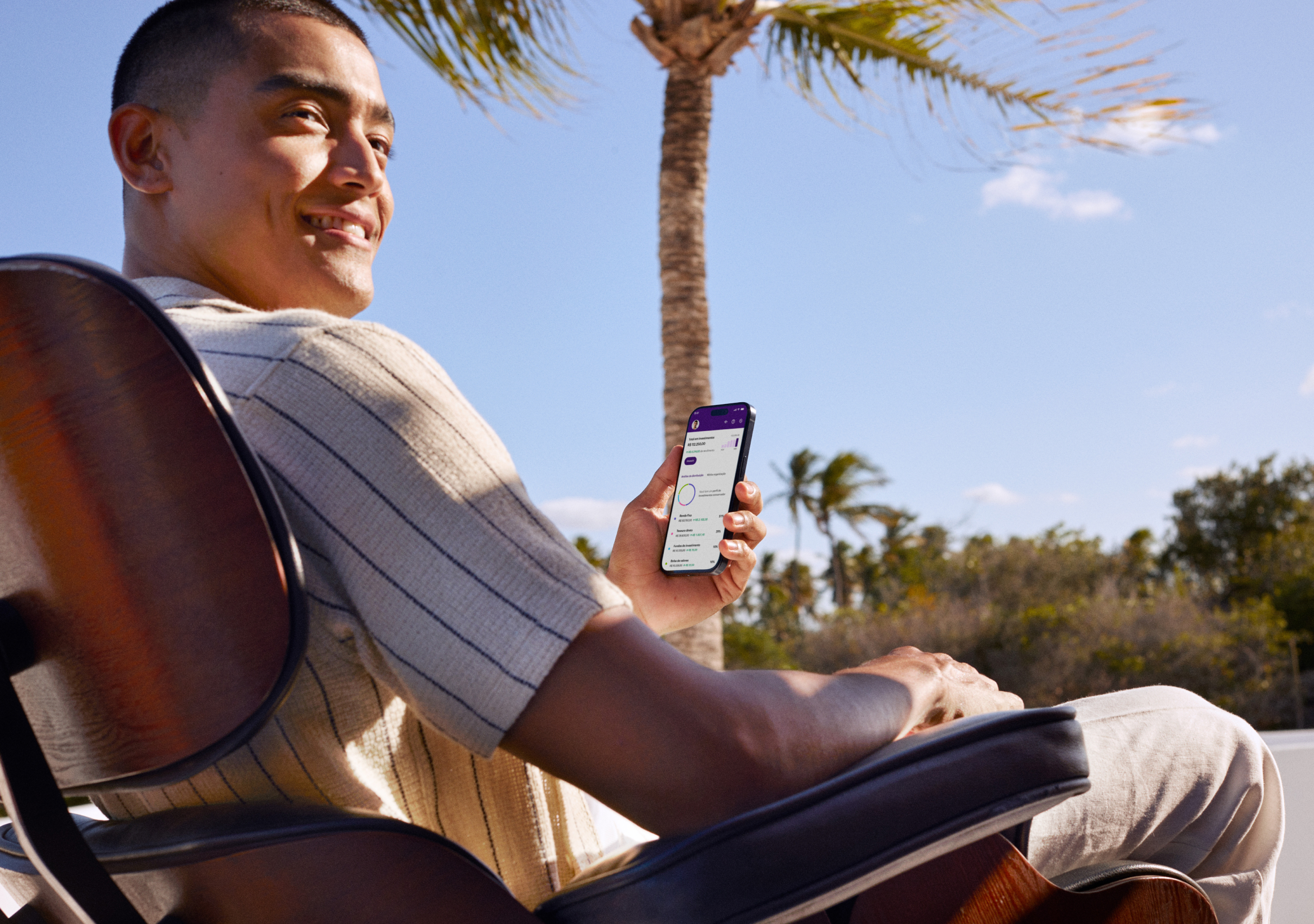 A imagem mostra um homem sorridente sentado em uma cadeira ao ar livre, segurando um smartphone com o aplicativo do Nubank aberto na tela.