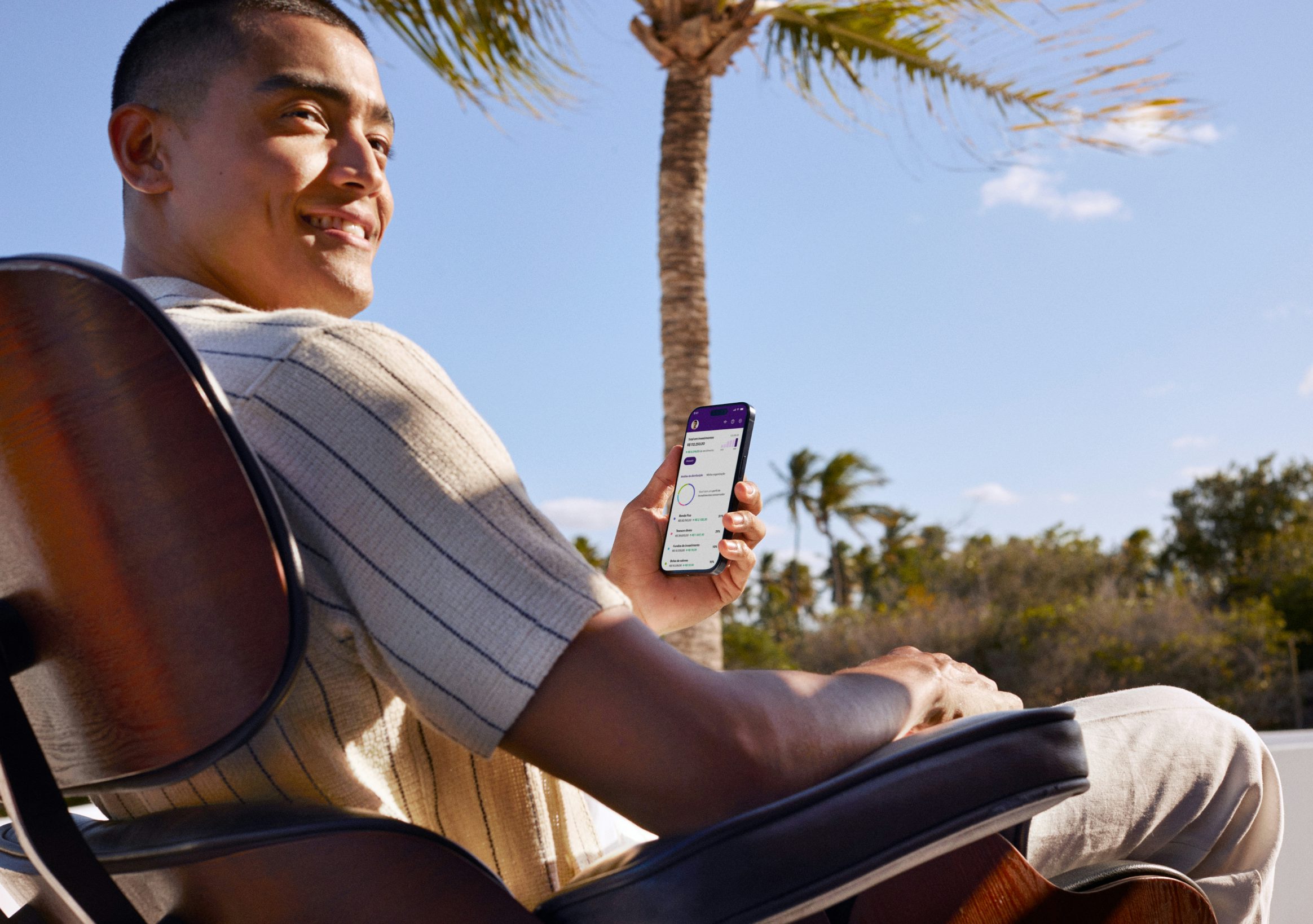 A imagem mostra um homem sorridente sentado em uma cadeira ao ar livre, segurando um smartphone com o aplicativo do Nubank aberto na tela.
