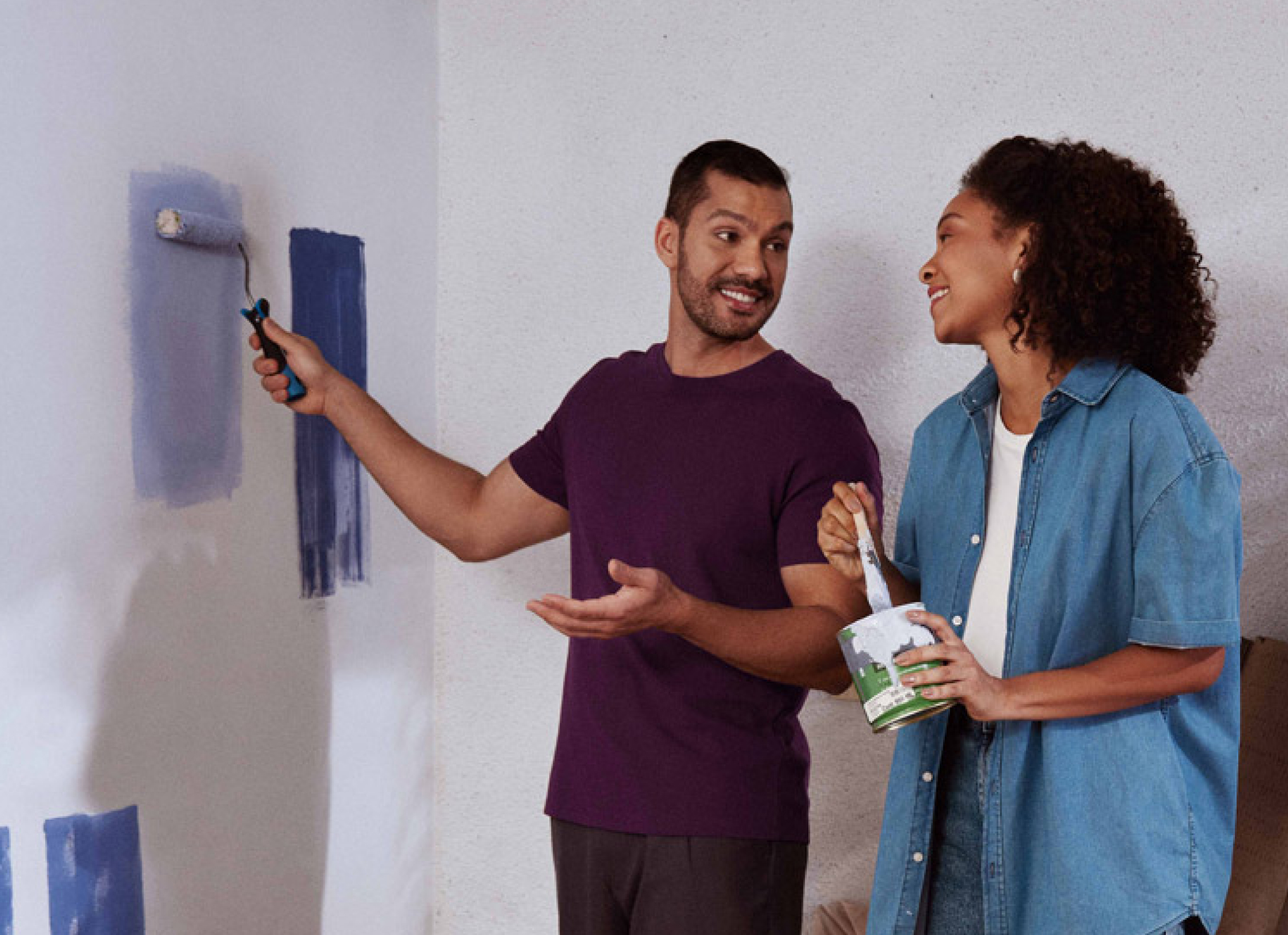 Homem em pé, passa com sua mão direita um rolo de tinta azul em uma parede branca, enquanto olha para a esquerda e conversa com uma mulher negra segurando com a mão esquerda uma lata de tinta branca e com a direita um pincel.