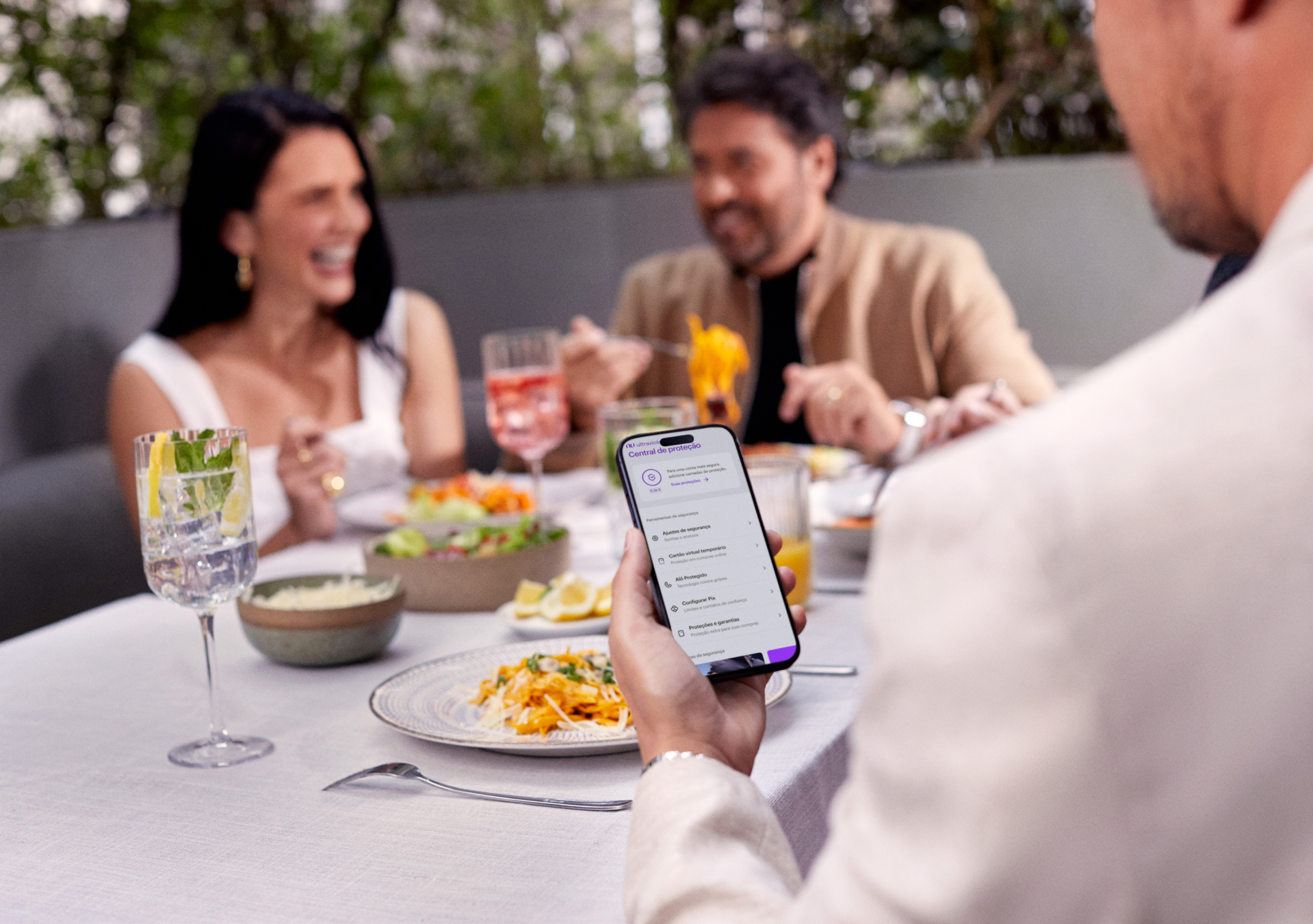 Homem de costas olha para o celular, que está em sua mão esquerda sobre uma mesa com drinks e comidas. Ele está checando a Central de Proteção Ultravioleta, no App do Nu. Ao fundo e de frente para ele, um casal rindo enquanto almoçam.