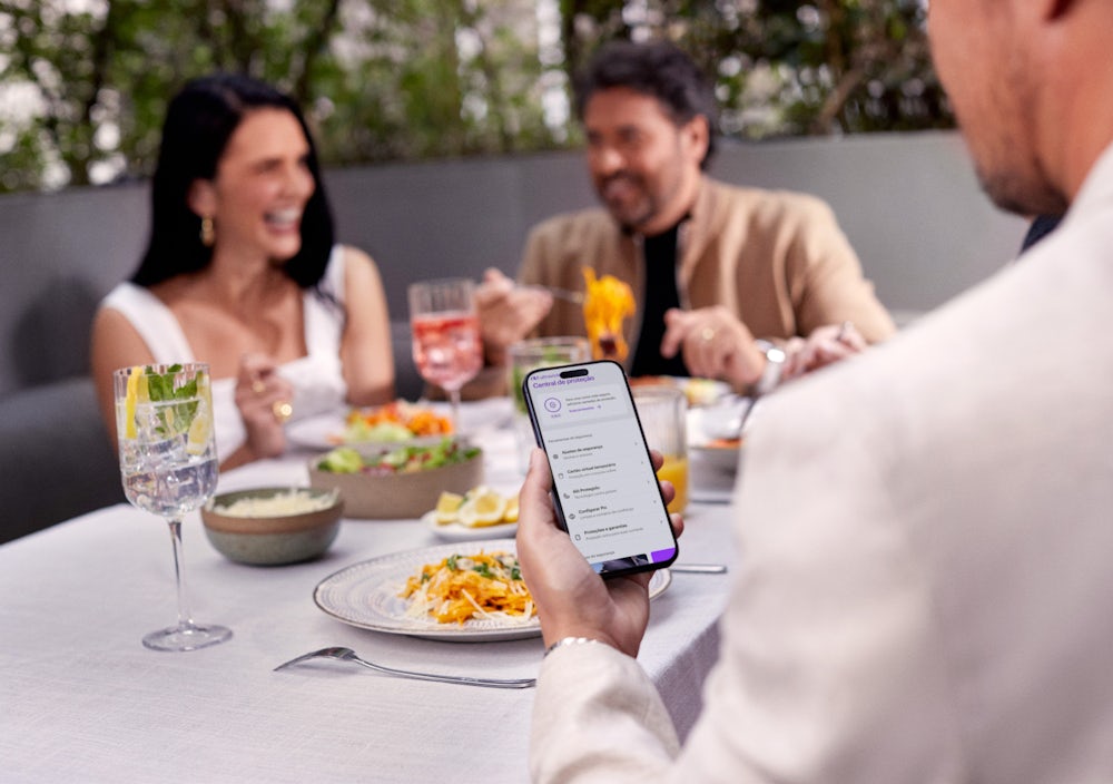 Homem de costas olha para o celular, que está em sua mão esquerda sobre uma mesa com drinks e comidas. Ele está checando a Central de Proteção Ultravioleta, no App do Nu. Ao fundo e de frente para ele, um casal rindo enquanto almoçam.