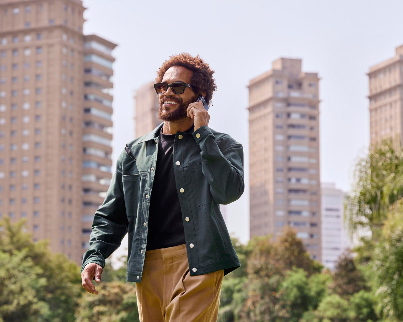 Homem sorrindo enquanto fala no celular passeando pela cidade. Ele está segurando o celular com a mão esquerda e, ao fundo, prédios e algumas árvores.