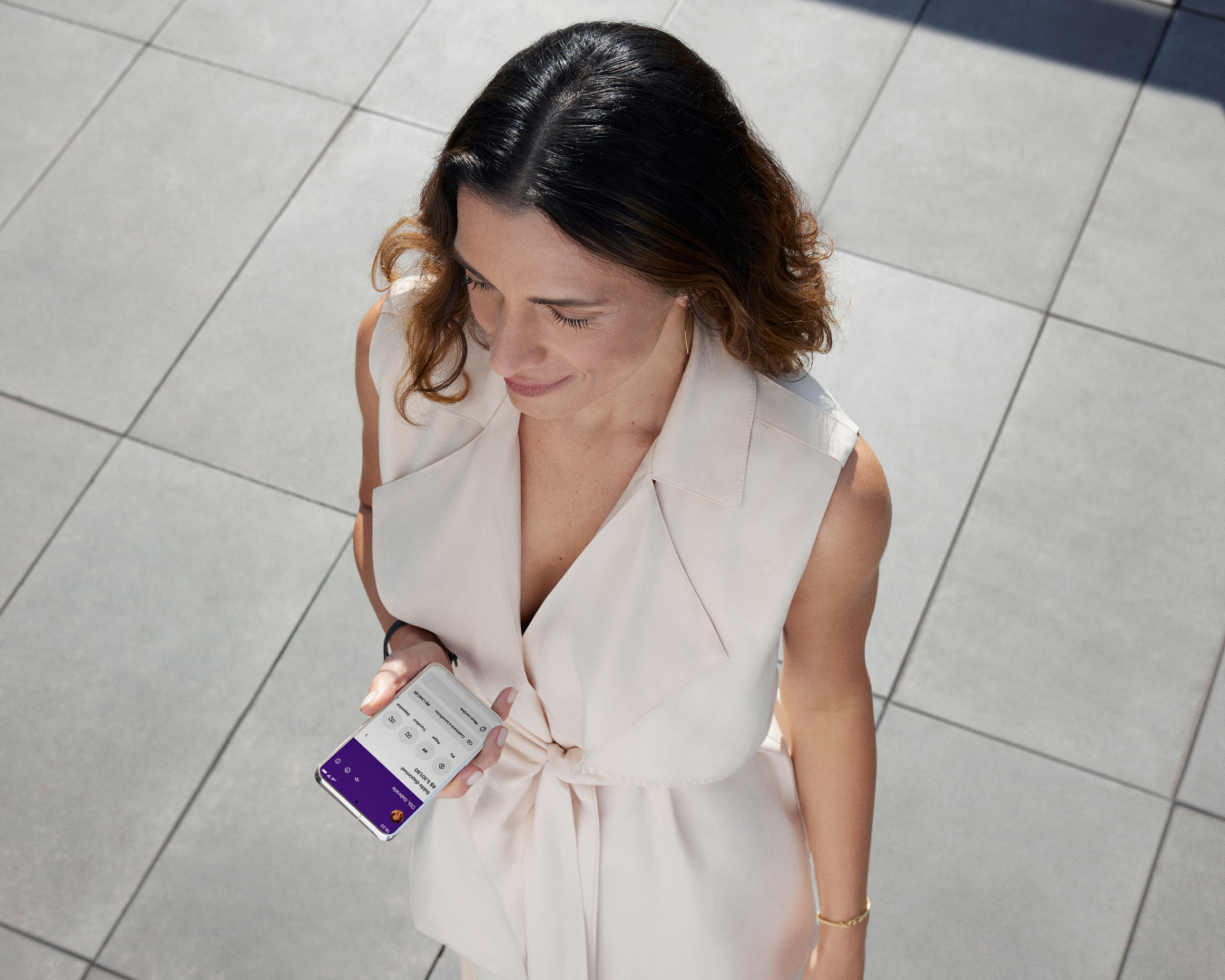 Vista de cima mostra uma mulher branca segurando o celular com a mão esquerda e olhando para frente. Na tela do celular, a sua conta Nu.
