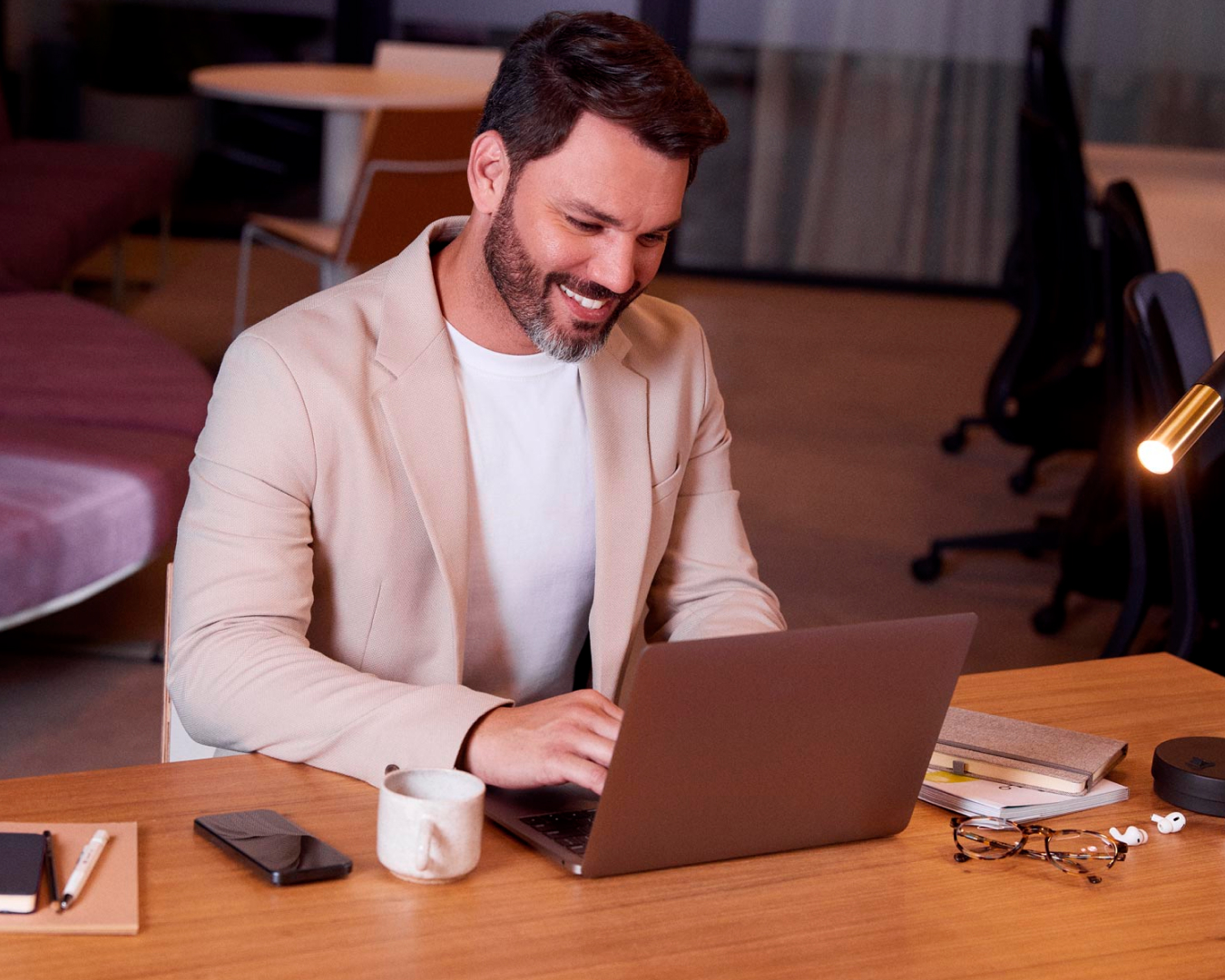 Homem branco sorrindo na sala de trabalho, enquanto faz um atendimento online por meio do notebook que está em cima da mesa com alguns itens de escritório, seu celular e uma luminária.