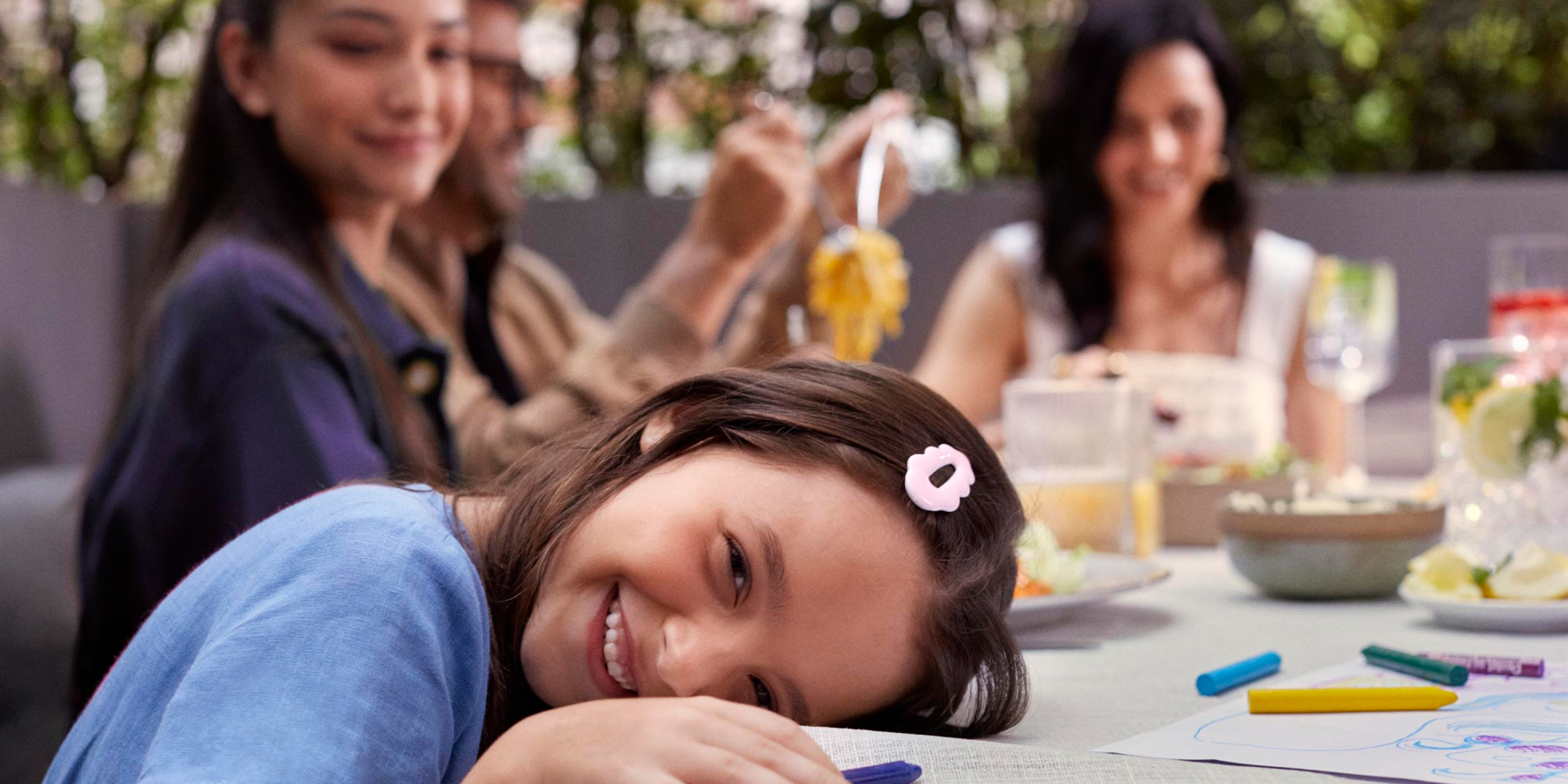 Em primeiro plano, uma criança está com a cabeça apoiada na mesa e com giz de cera na mão. Ao fundo, uma menina mais velha olha para criança, enquanto um casal almoça.