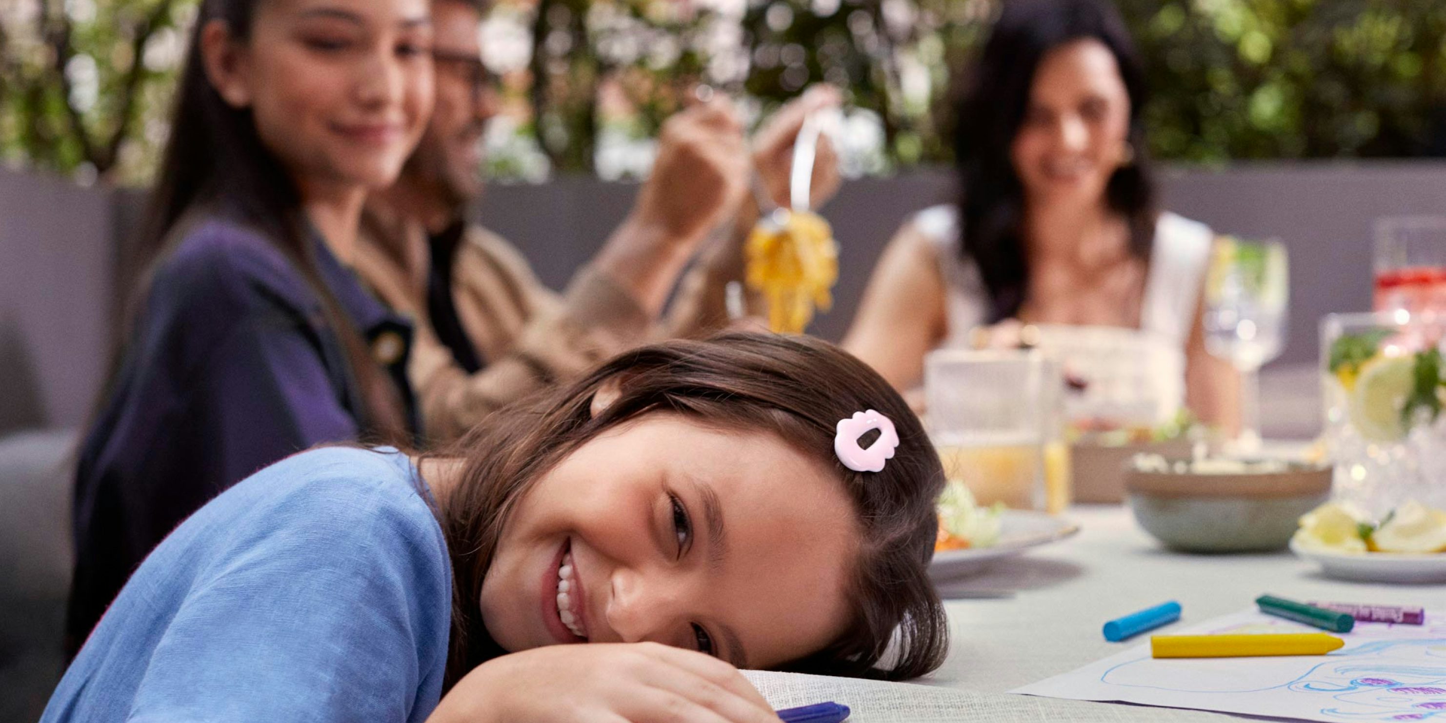 Em primeiro plano, uma criança está com a cabeça apoiada na mesa e com giz de cera na mão. Ao fundo, uma menina mais velha olha para criança, enquanto um casal almoça.
