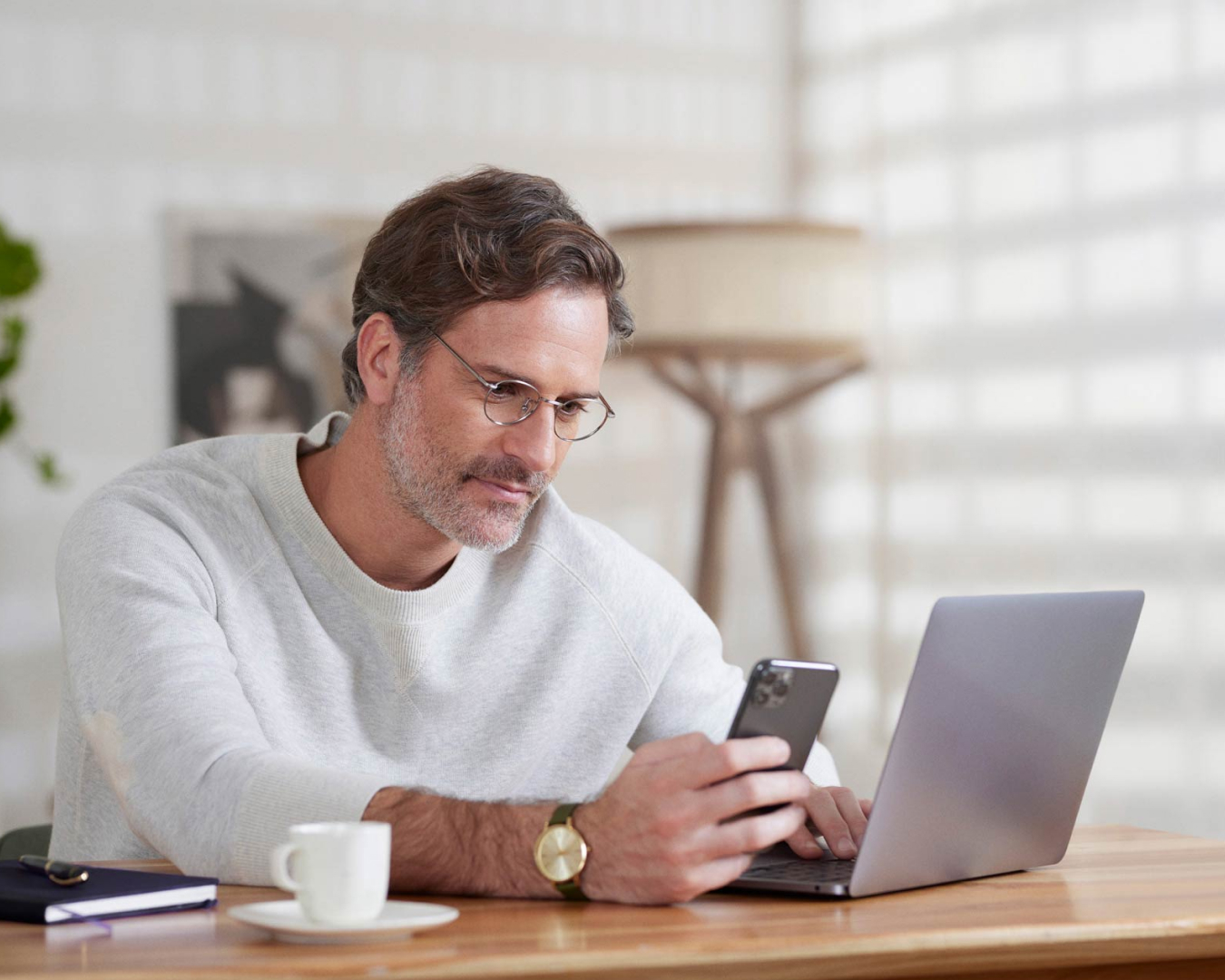 Homem de óculos sentado em um escritório olhando para a tela do celular que está em uma de suas mãos. A outra mão está em cima do teclado do notebook. Na mesa, uma xícara de café, uma agenda e uma caneta.