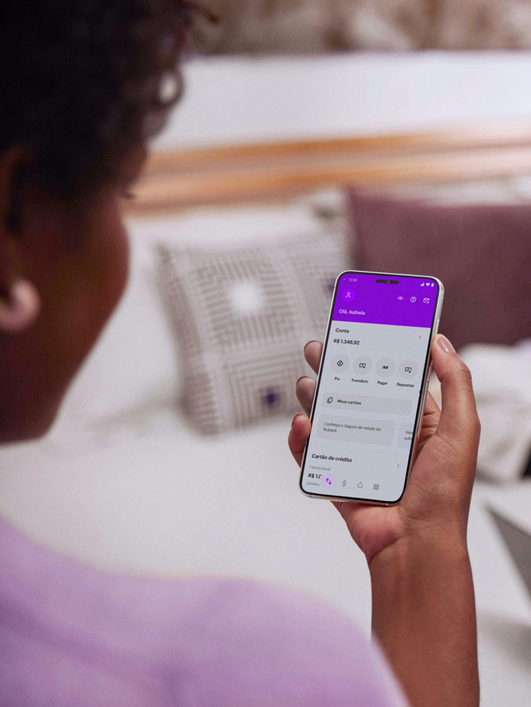 Vista das costas de uma mulher negra sentada com um celular na mão direita e visualizando sua conta no aplicativo do Nubank.