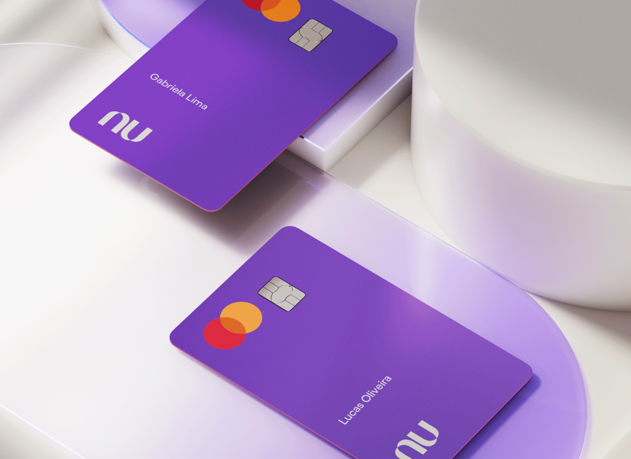 Dois cartões do Nubank vistos de cima postos sob superfícies geométricas circulares. Os cartões estão com a face principal virada para cima, com o logo da bandeira Mastercard, chip e nomes dos clientes.