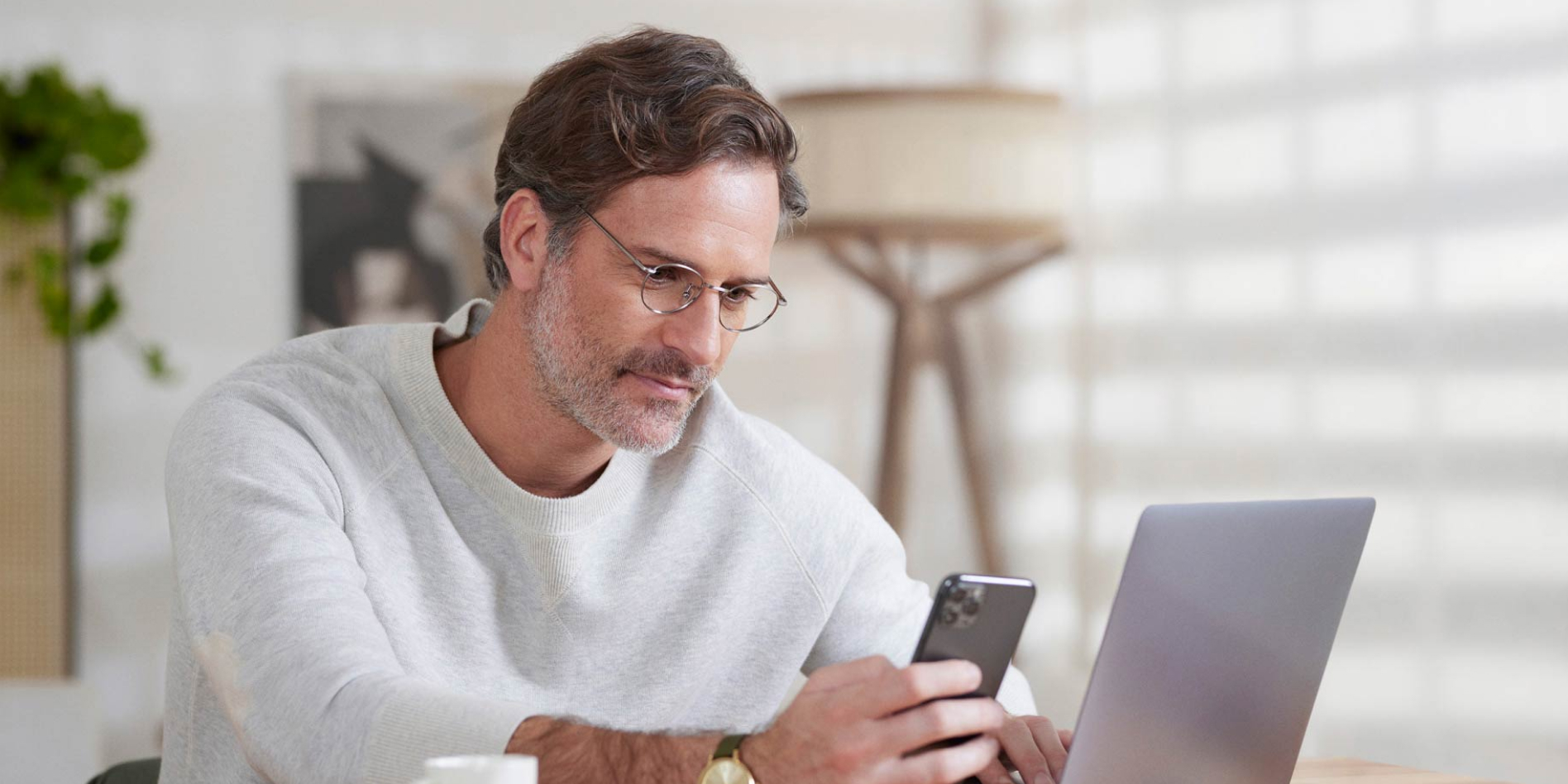 Homem de óculos sentado em um escritório olhando para a tela do celular que está em uma de suas mãos. A outra mão está em cima do teclado do notebook. Na mesa, uma xícara de café, uma agenda e uma caneta.