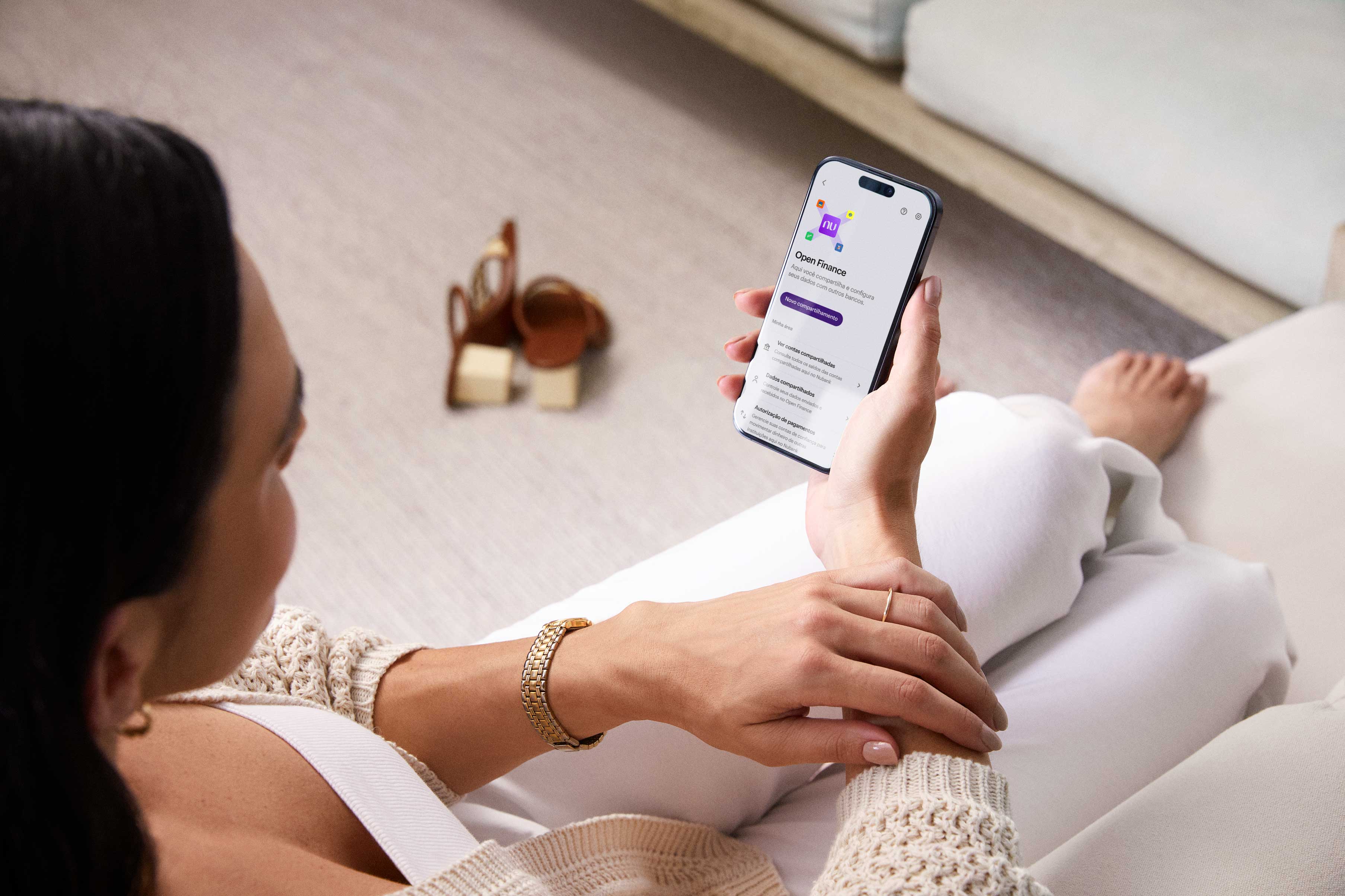 Uma mulher branca de cabelos pretos sentada e com os pés em um sofá branco segura o celular que está em evidência com a mão direita e visualiza a seção Open Finance no aplicativo do Nubank. No seu braço esquerdo, um relógio e um anel fino. Ao fundo, uma sandália marrom no chão.