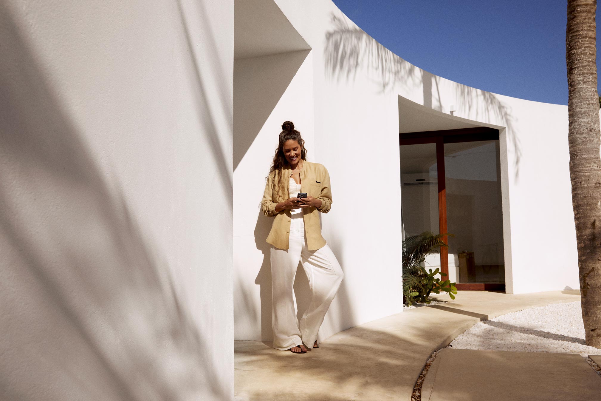 Mulher encostada em uma parede branca com a sombra de coqueiros em ambiente externo. Ela está sorrindo e olhando para o smartphone que está em suas mãos.