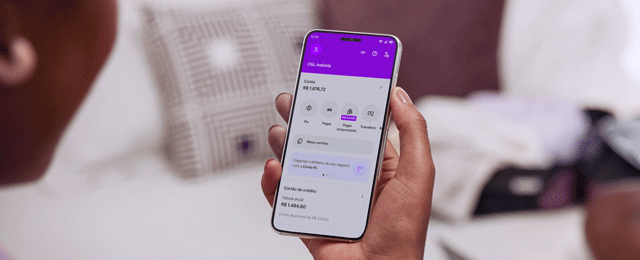 Vista das costas de uma mulher negra sentada com um celular na mão direita e visualizando sua conta no aplicativo do Nubank.