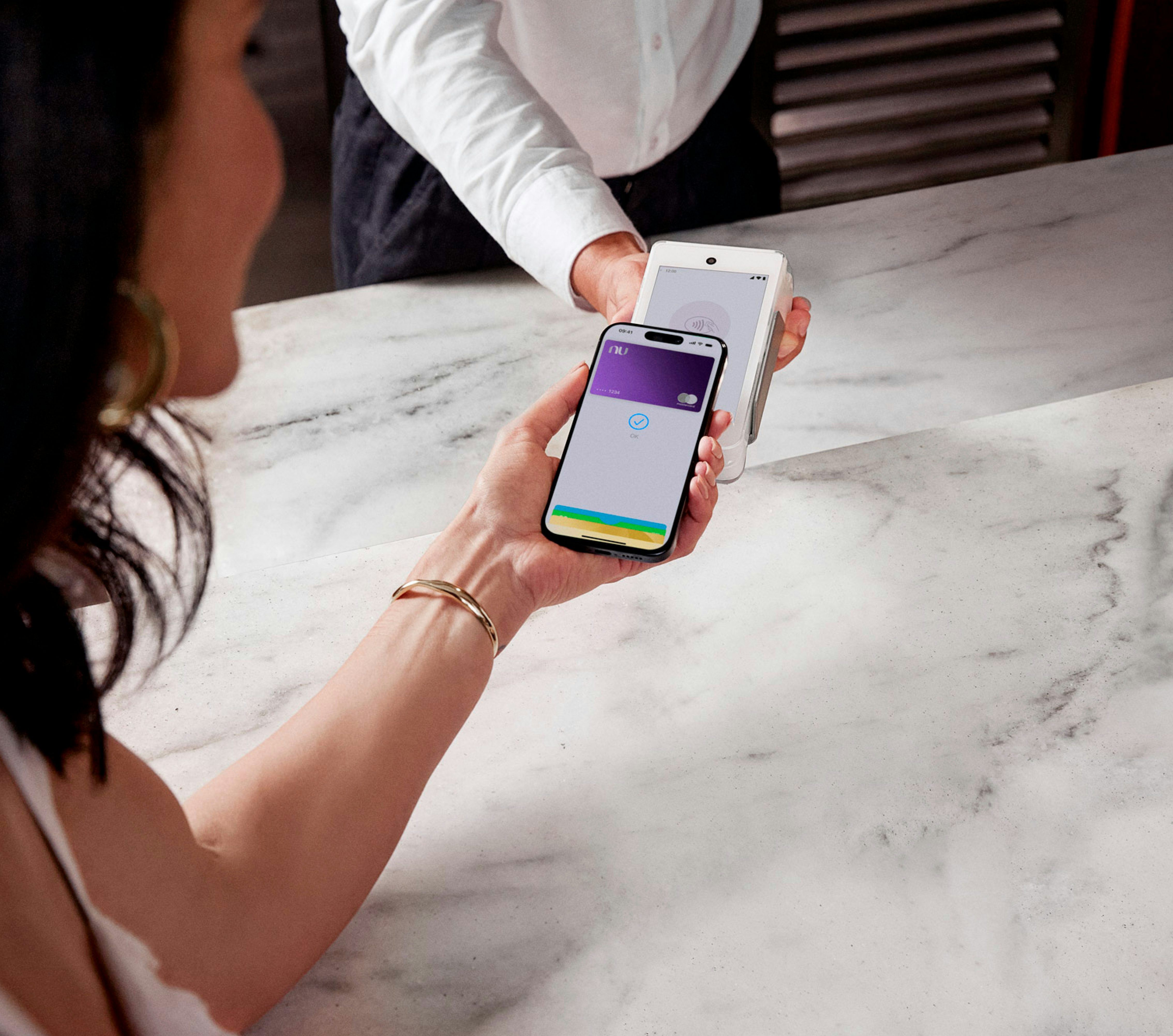 Vista de cima do perfil de uma mulher fazendo pagamento por aproximação a mão de um homem no outro lado da mesa segura a maquininha.