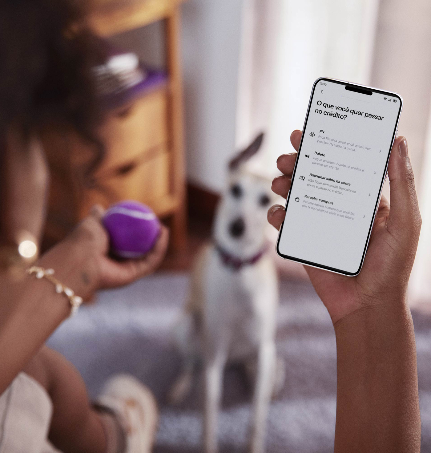 Vista das costas de uma mulher negra sentada, com um celular na mão direita e visualizando sua conta no aplicativo do Nubank, com sua mão esquerda segura uma bolinha de tênis roxa, ao fundo e desfocado um cachorro de pequeno porte sentado no chão olhando para a bolinha.