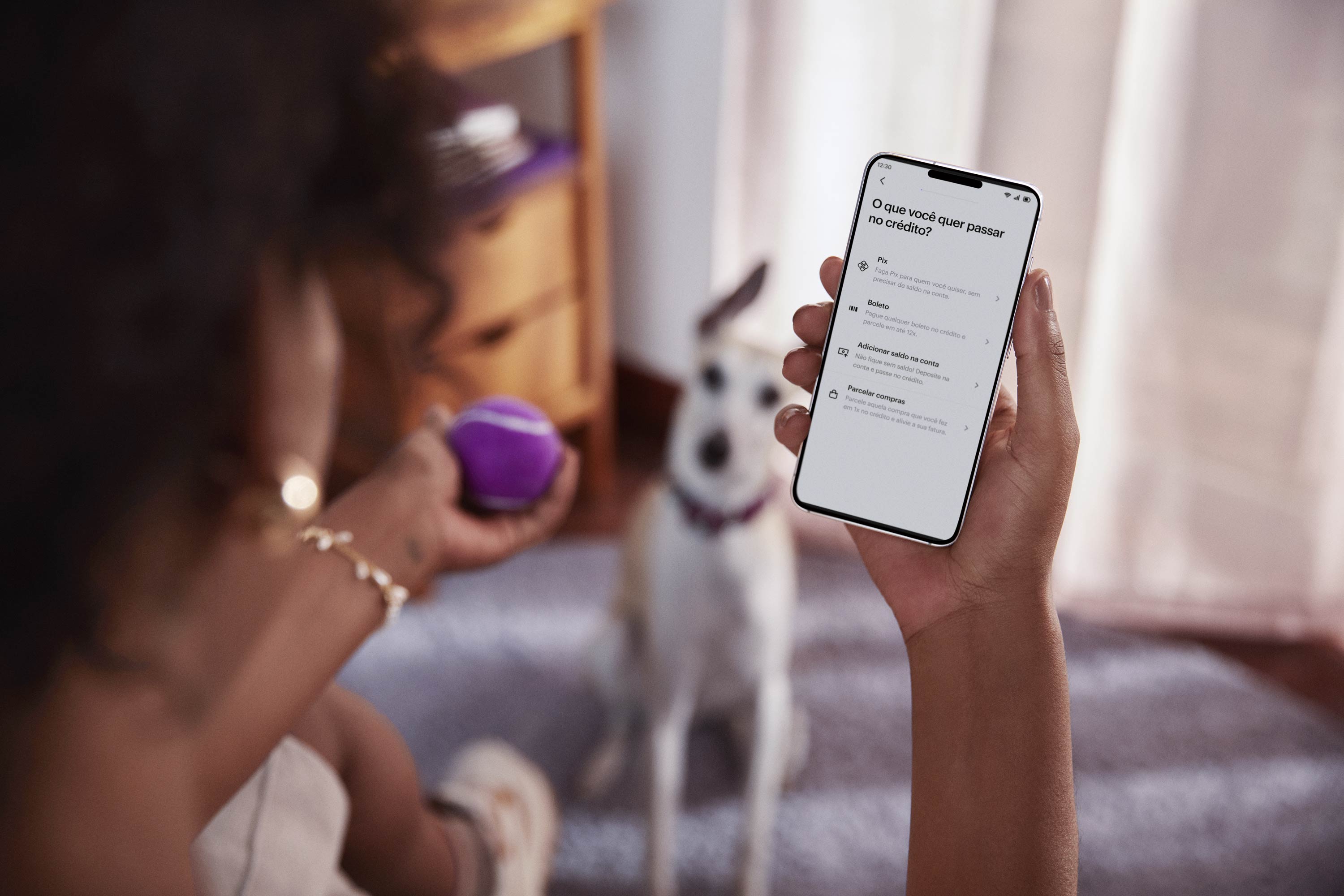 Vista das costas de uma mulher negra sentada, com um celular na mão direita e visualizando sua conta no aplicativo do Nubank, com sua mão esquerda segura uma bolinha de tênis roxa, ao fundo e desfocado um cachorro de pequeno porte sentado no chão olhando para a bolinha.