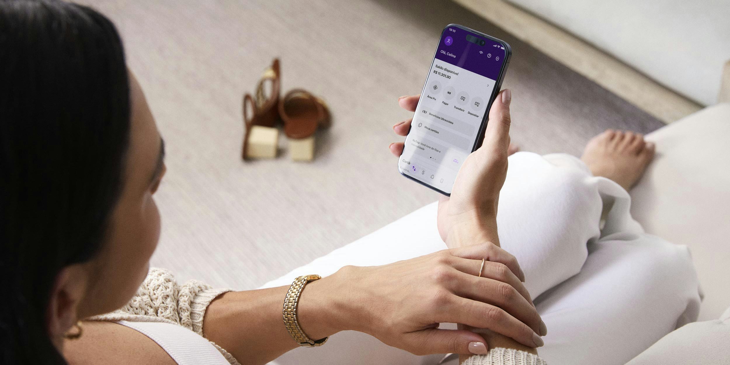 Uma mulher branca de cabelos pretos sentada e com os pés em um sofá branco, segurando o celular e visualizando sua conta no aplicativo do Nubank. Ao fundo, sua sandália marrom está no chão.