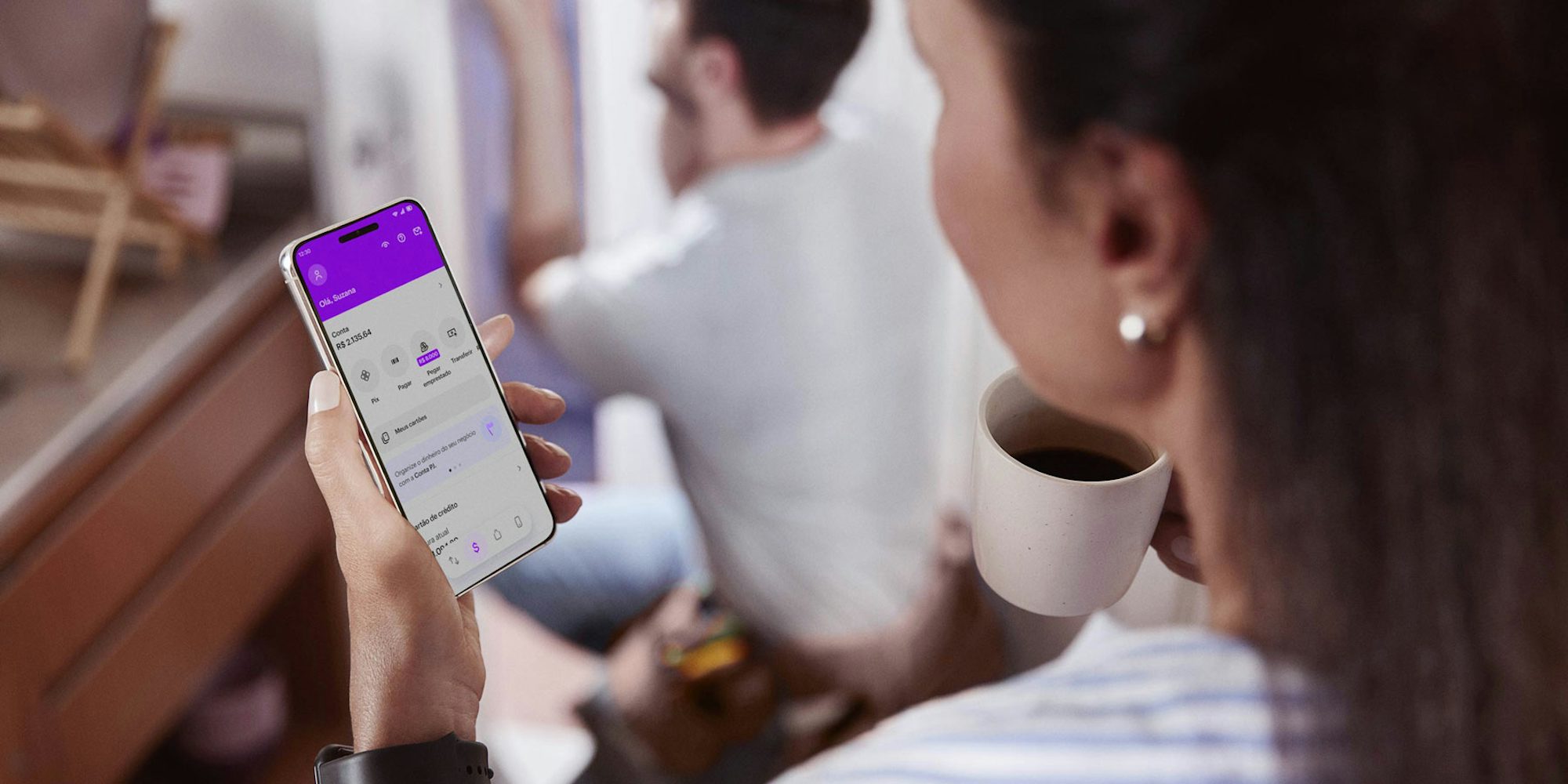 Vista das costas de uma mulher branca em pé em uma cozinha, com um celular na mão esquerda e visualizando sua conta no aplicativo do Nubank e segurando uma xícara de café com a mão direita, ao fundo e desfocado um homem branco de joelhos consertando uma porta.