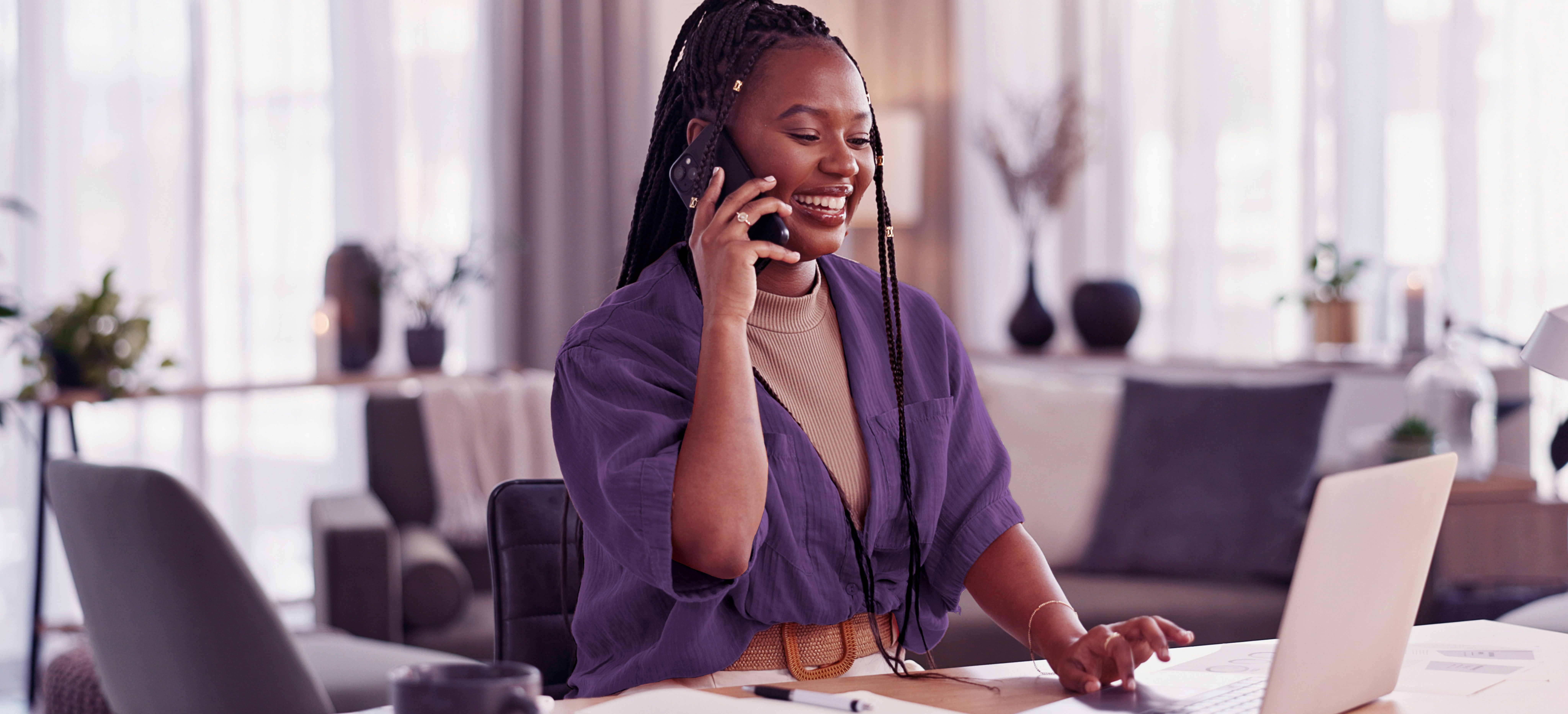 Imagem mostra uma mulher negra de cabelos trançados sorrindo enquanto fala ao telefone. Ela veste uma camisa roxa sobre uma blusa bege, uma calça clara e um cinto marrom. Ela está em uma mesa de escritório usando um lap top.