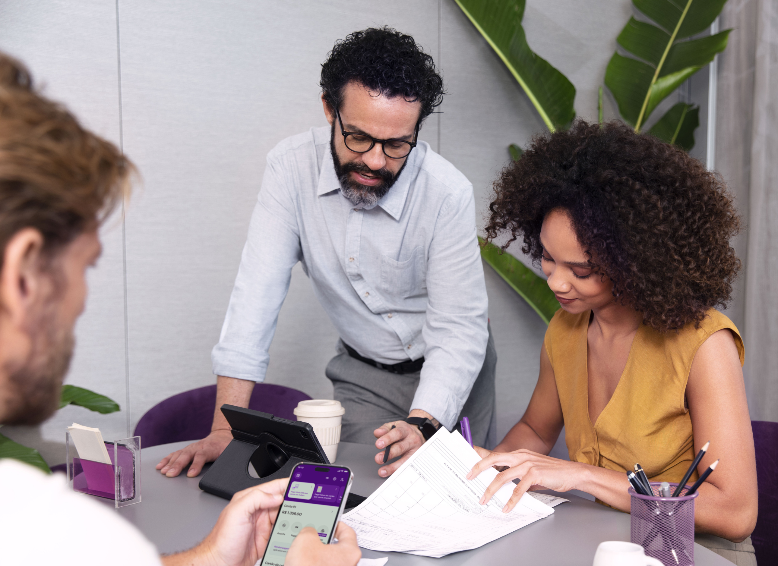Homem de costas olha o celular em uma mesa de escritório com outras duas pessoas: um homem branco de barba e cabelos pretos vestindo uma calça social cinza escuro e uma camisa cinza claro; e uma mulher negra de cabelos pretos usando um vestido amarelo. A mulher assina alguns papeis enquanto um dos homens a observa. Na mesa é possível ver um aparelho eletrónico para videochamadas, canetas, um porta cartões e um copo. Ao fundo, existe uma planta.