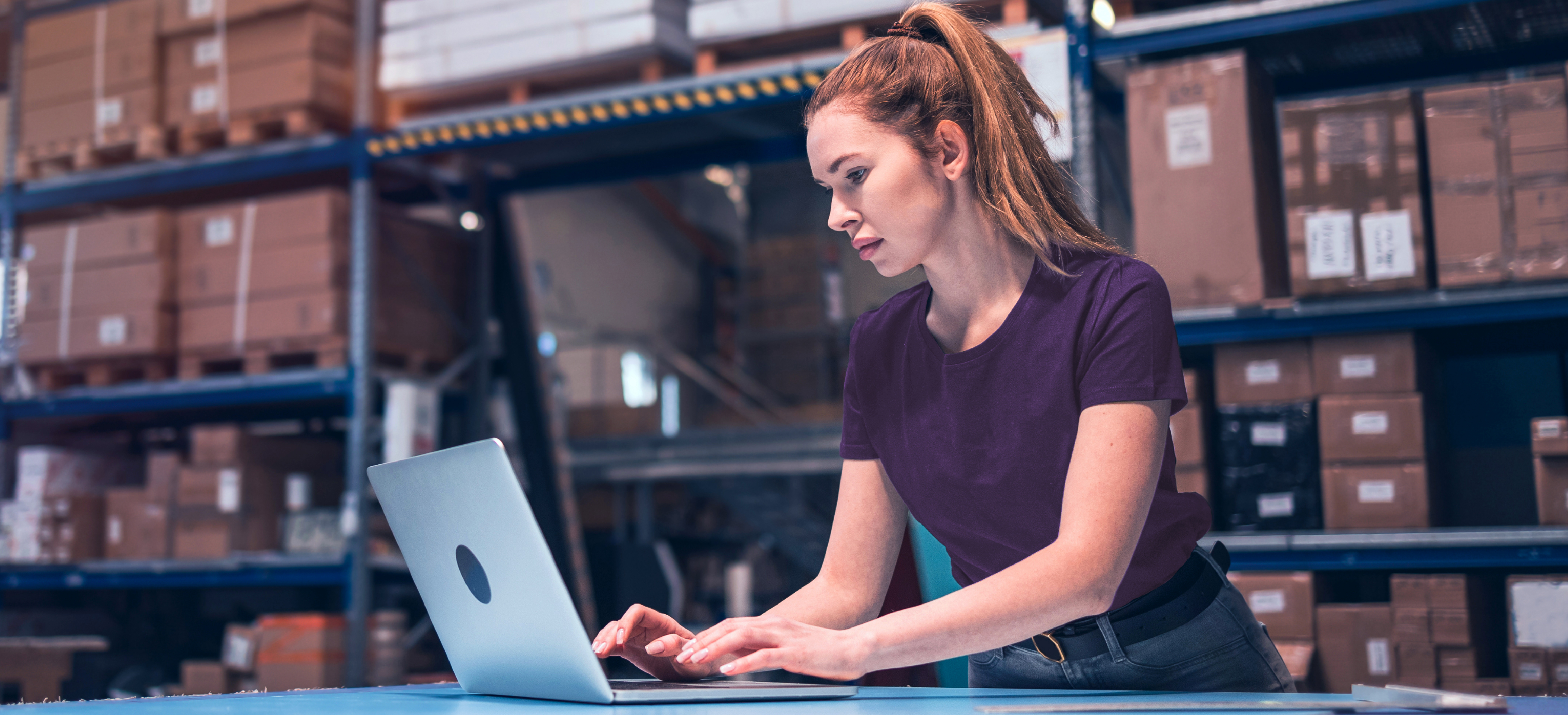 Uma mulher branca de cabelos longos usa um lap top em cima de uma bancada. Ele veste uma camiseta roxa e calça jeans. Atrás dela existe uma grande prateleira de depósito cheia de caixas.