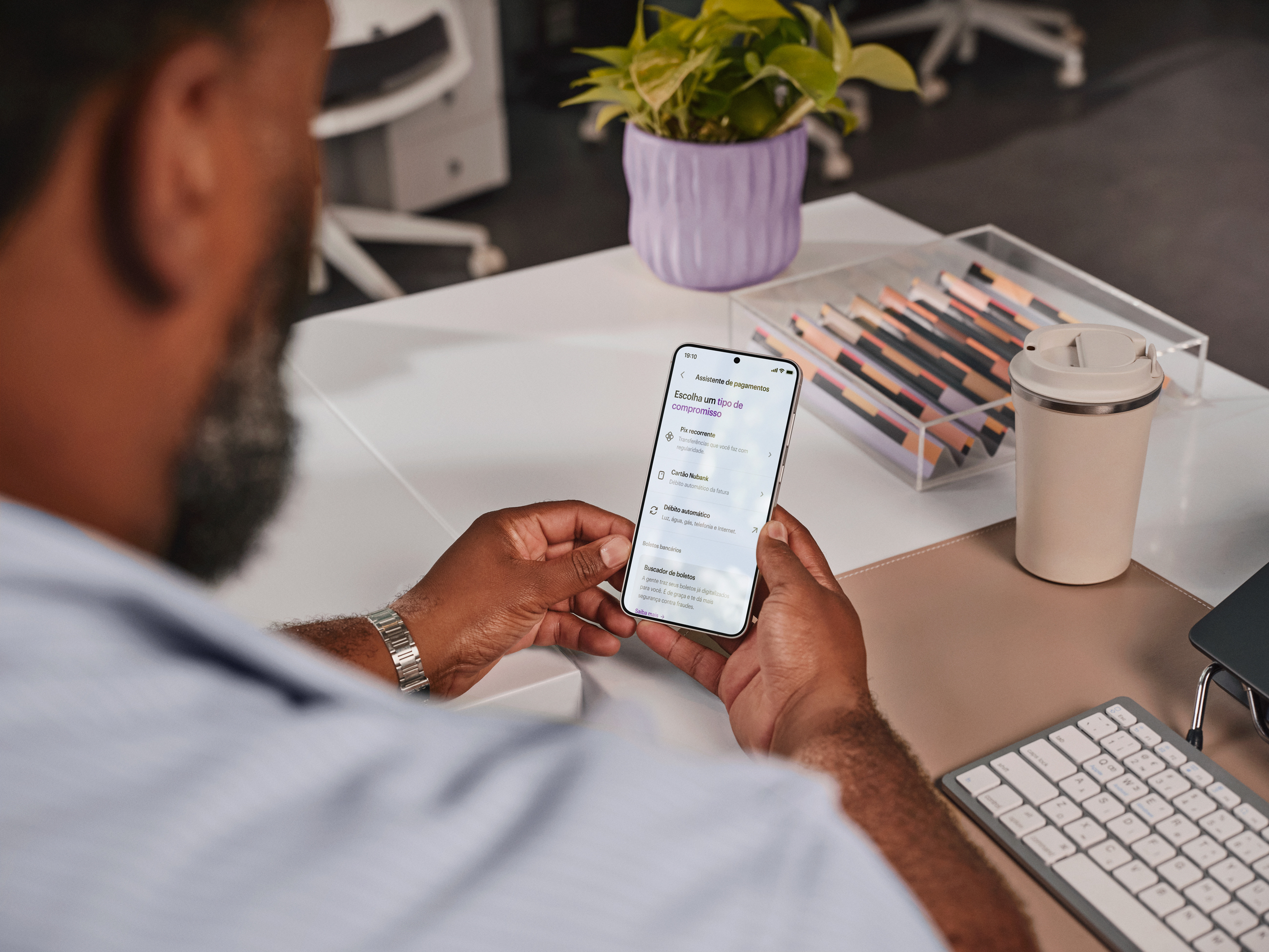 Homem de costas olha o celular em uma mesa de escritório.
