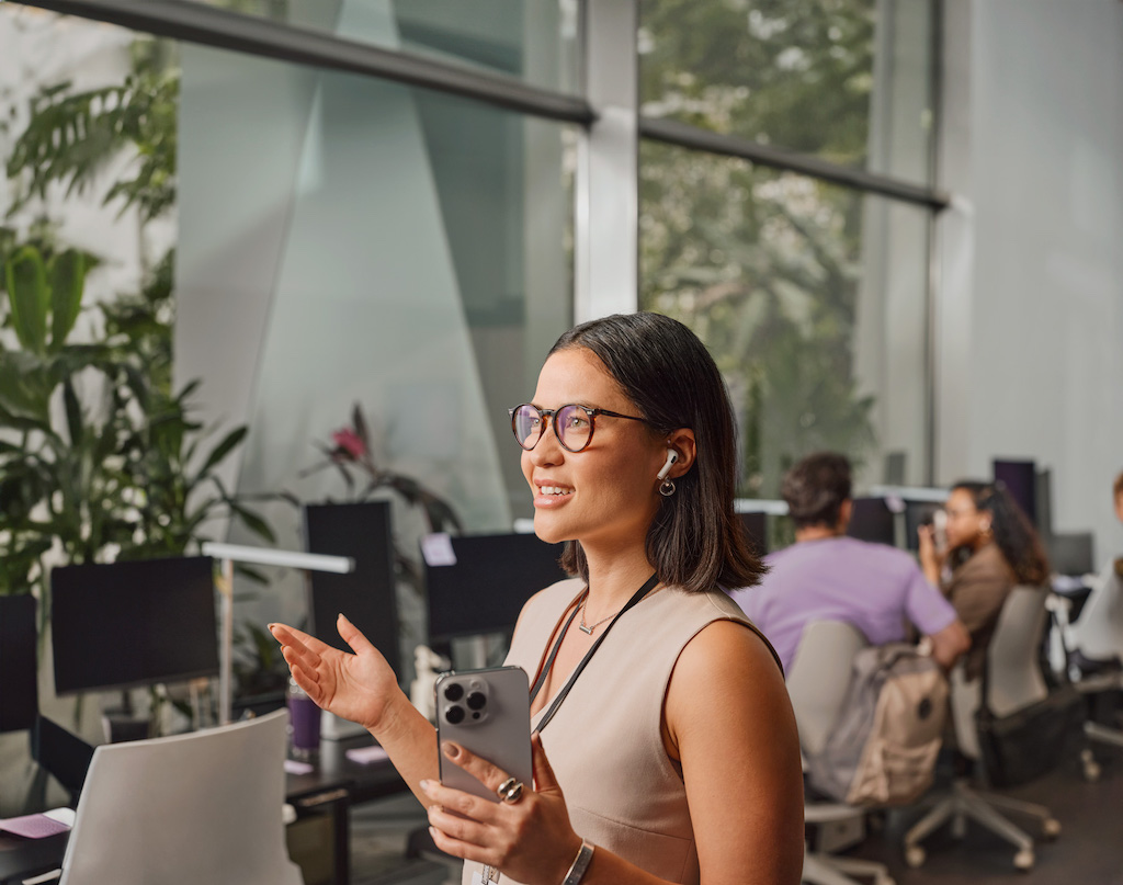 Imagem mostra uma mulher branca, de óculos e cabelos lisos na altura dos ombros, gesticulando com as mãos enquanto sorri e olha para frente. Ela segura um celular na mão esquerda. Ao fundo, pessoas trabalham em mesas de escritório.