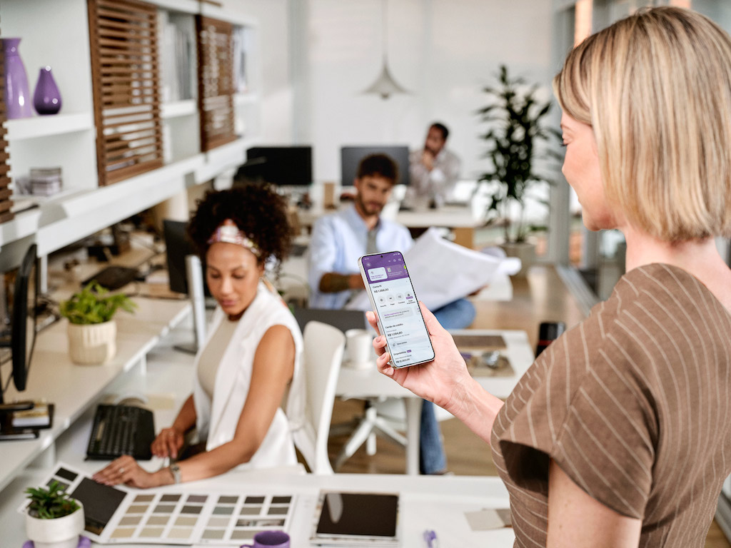 Escritório com móveis brancos. Em primeiro plano está uma mulher branca segurando um celular na mão direita. Na tela, está o app da conta Nu Empresas. Em segundo plano, uma mulher negra trabalha em uma mesa de escritório. Ela veste uma roupa branca. Outras pessoas trabalham em mesas ao fundo.