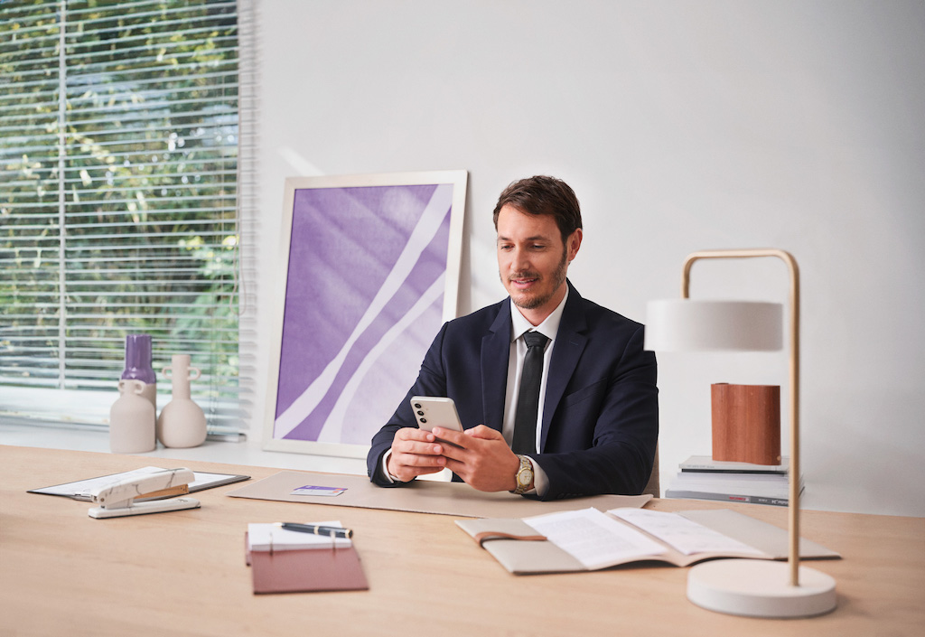 Homem branco vestindo terno olha o celular sentado em uma mesa de escritório