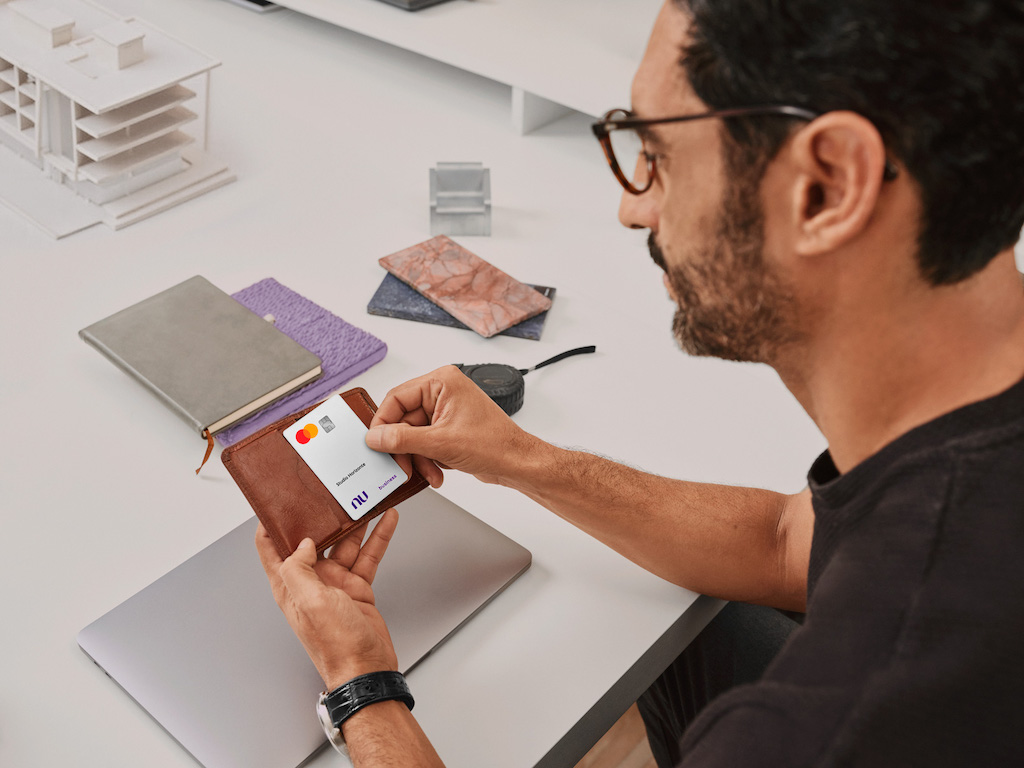 Homem branco sentado em uma mesa de escritório segura uma carteira e um cartão Nu Empresas. Ele veste uma camiseta preta, usa óculos e tem barba e cabelos pretos. Na mesa branca está um notebook fechado e objetos de escritório como cadernos.