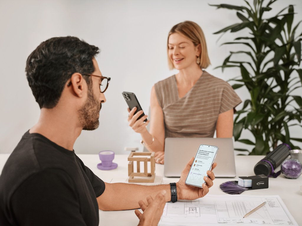 Em primeiro plano, um homem de barba e cabelos curtos, de óculos, usando uma camiseta preta olha para a tela do celular.  Ao fundo, uma mulher branca usando um vestido marrom olha para o celular. Eles estão sentados em uma mesa de escritório.