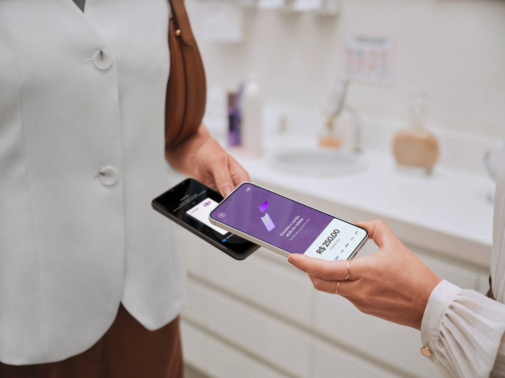 Imagem foca nas mãos de duas pessoas realizando uma transação de pagamento com os seus celulares. Em um dos celulares, é possível ver a tela do Tap to Pay do Nu Empresas.