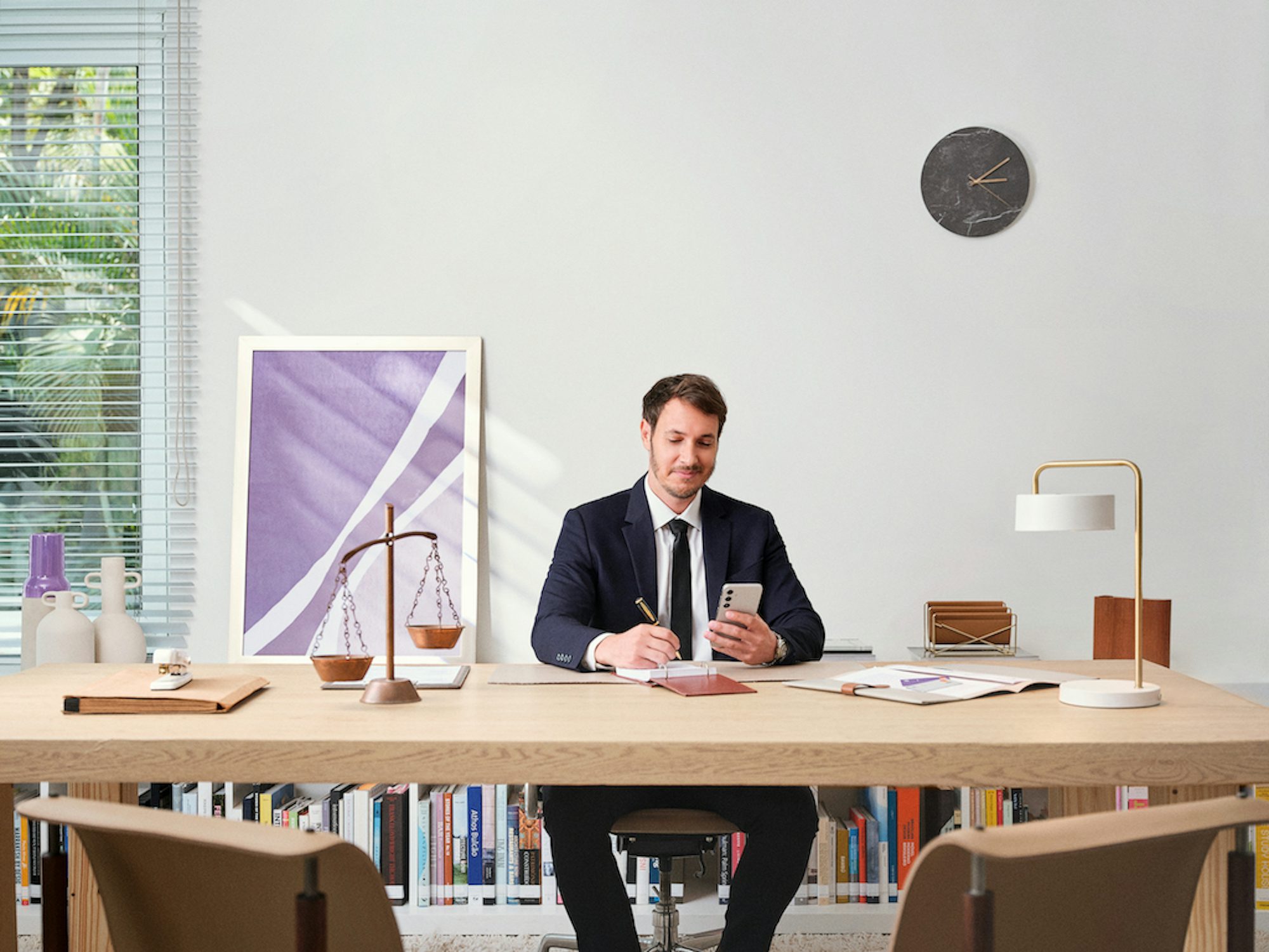 Homem branco vestindo terno olha o celular sentado em uma mesa de escritório