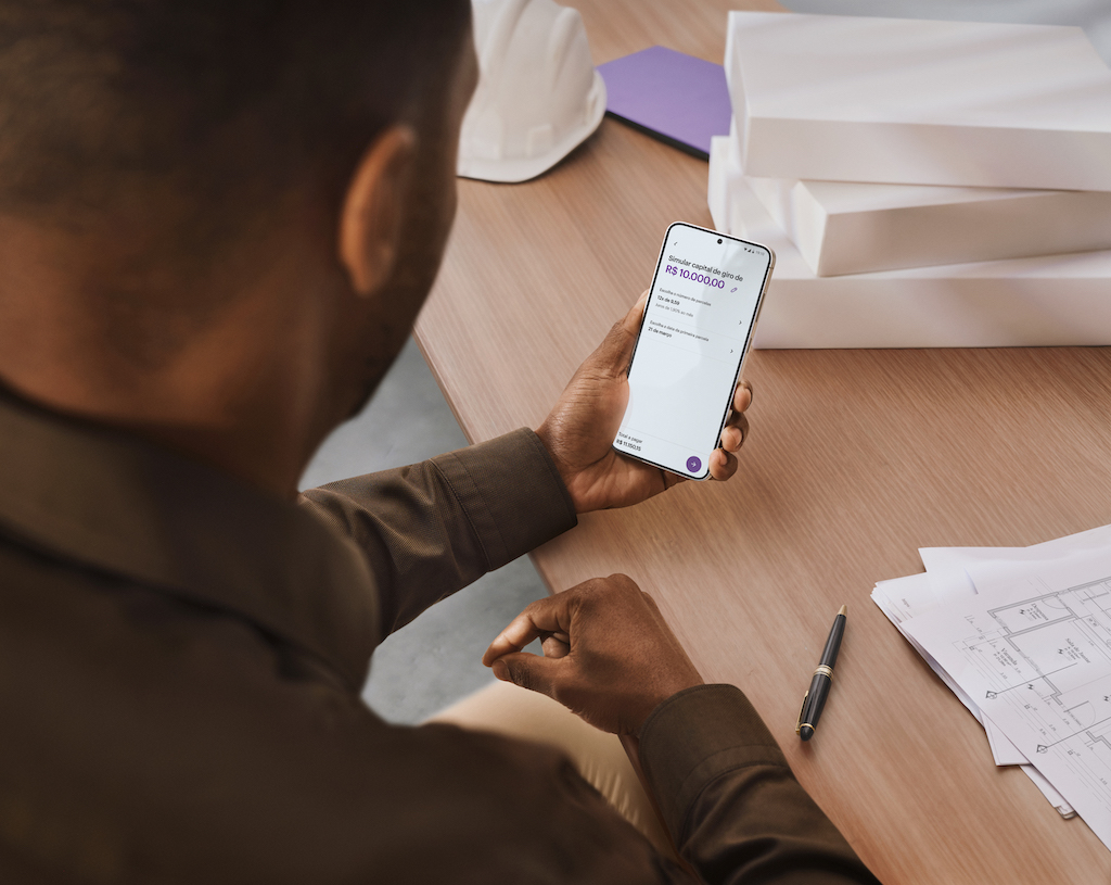 Homem sentado em uma mesa enquanto usa o celular. Ele usa uma camisa marrom. Na tela, é possível ver a função Capital de Giro do Nu Empresas.