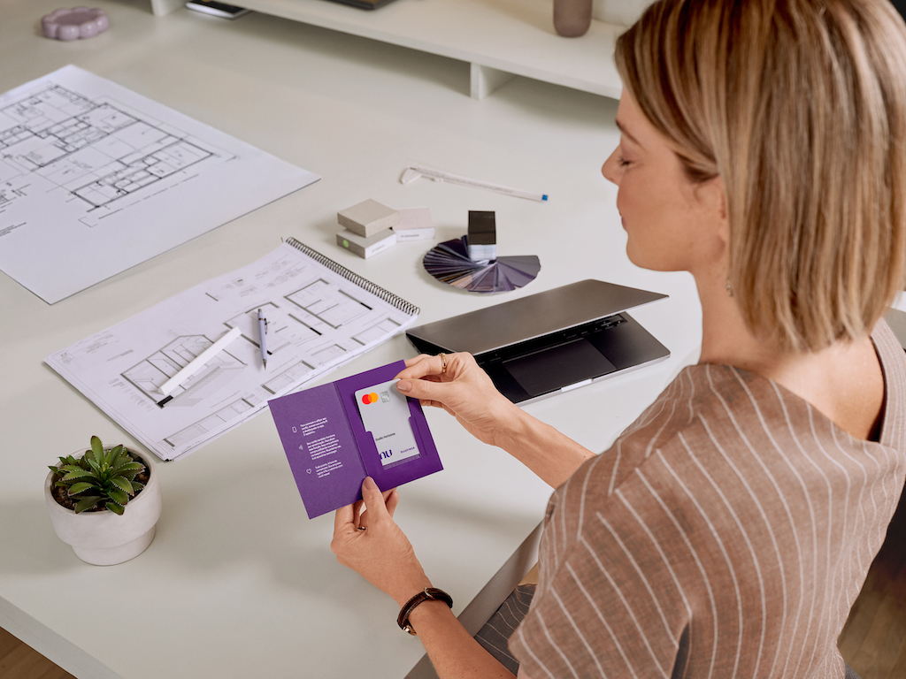 Imagem de uma mulher branca, de cabelos na altura do pescoço, segurando um cartão Nu Empresas. Ela usa um vestido marrom com listras brancas. Ela está em frente a uma mesa de escritório com papeis em cima, canetas e um notebook.